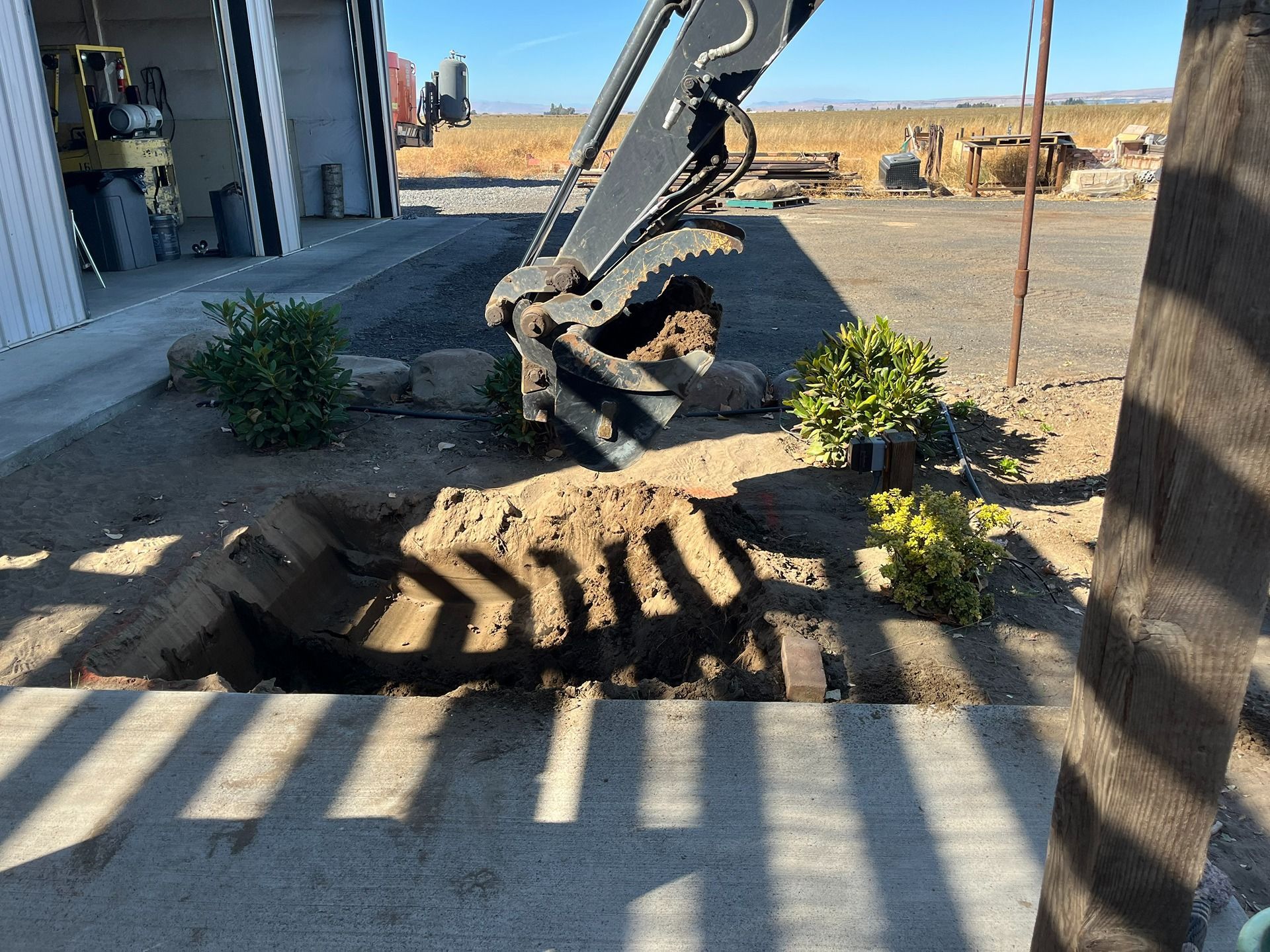An excavator arm hovering over a dug-out patch of dirt and soil near a building and some small bushes.