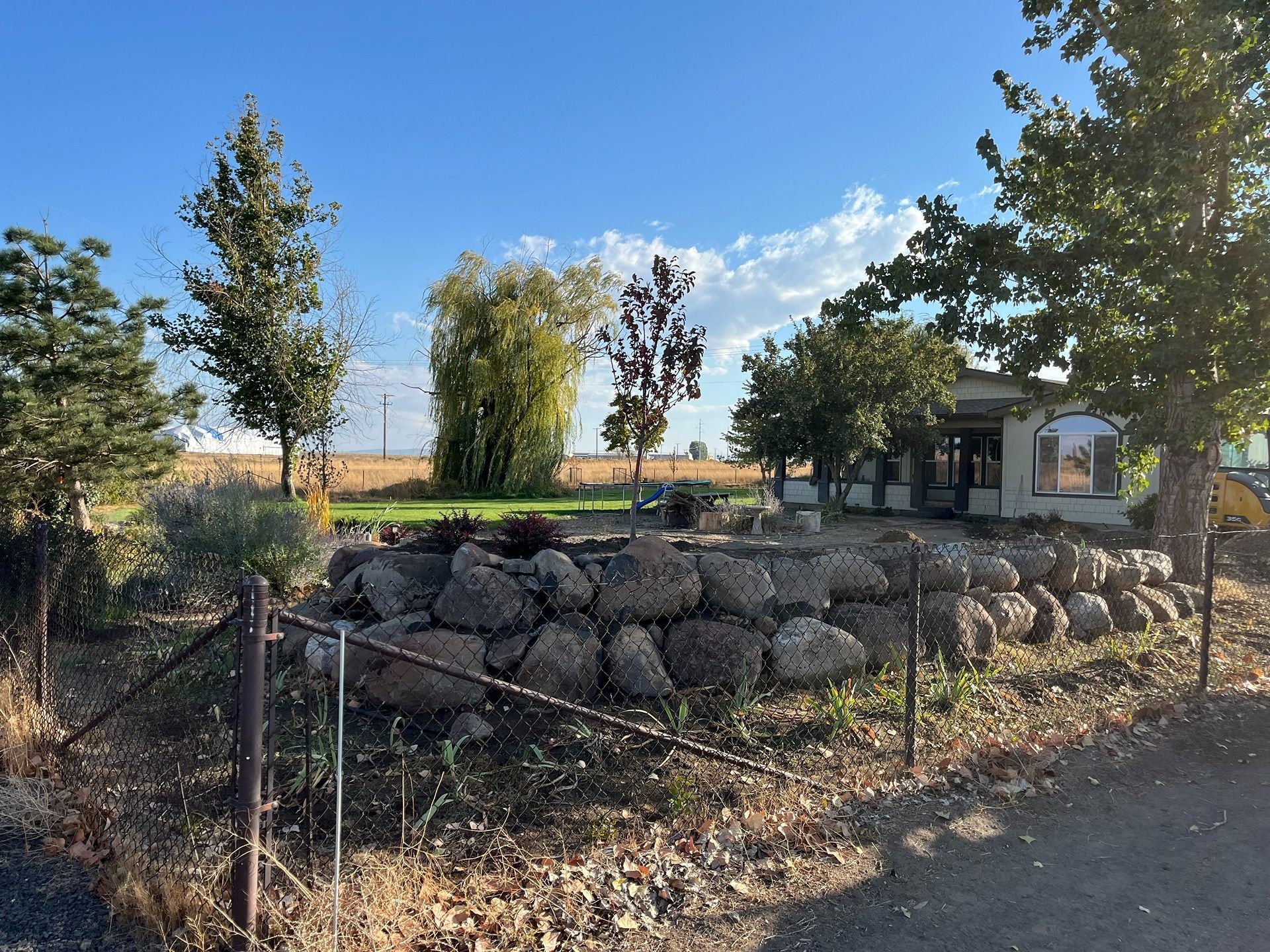 A large stone retaining wall sits in front of a house, surrounded by trees and a chain-link fence under a clear blue sky.