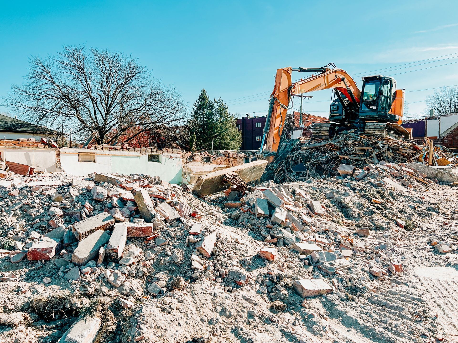 A yellow excavator clears a pile of brick and concrete rubble at a demolition site under a clear blue sky.