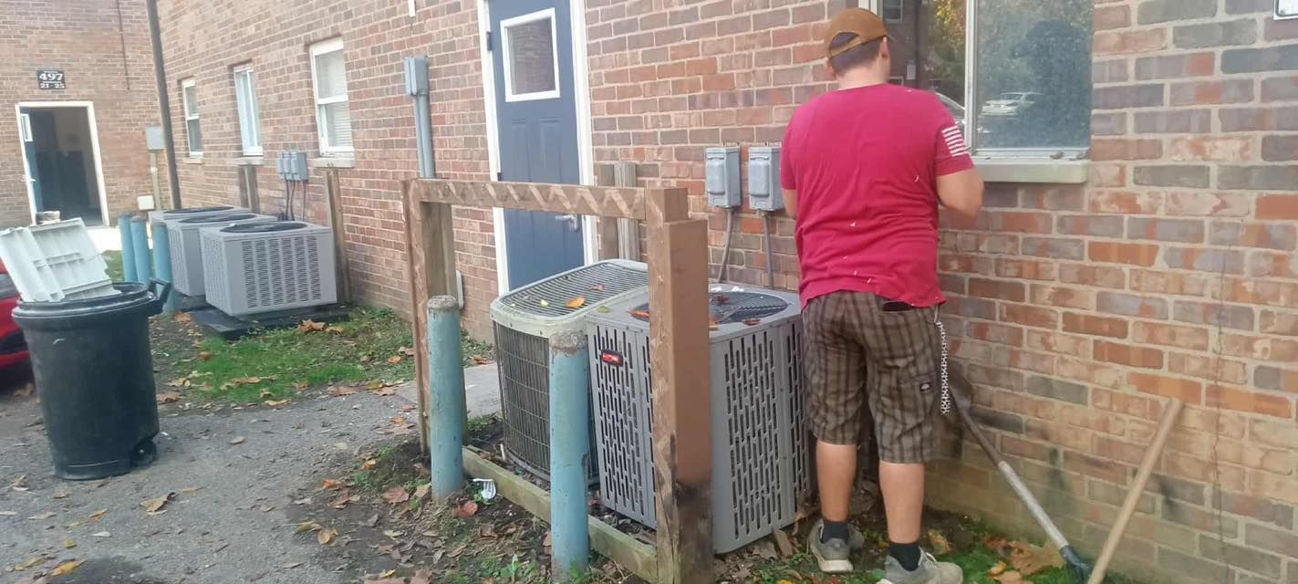 Person in a red shirt standing by a brick wall near utility units, working at a window.