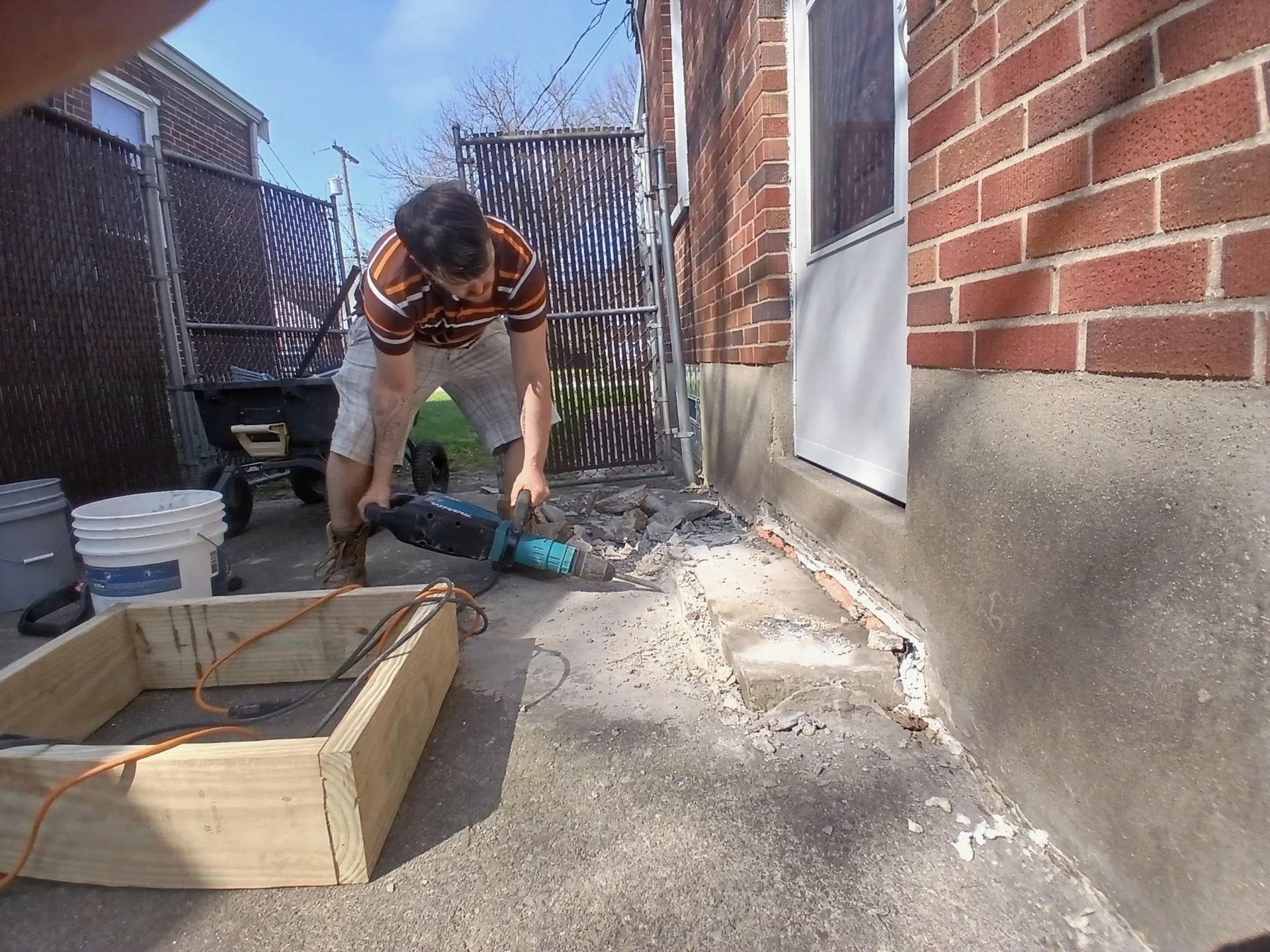Person kneels beside a small dog on a concrete patio next to a brick house and wooden planter box.