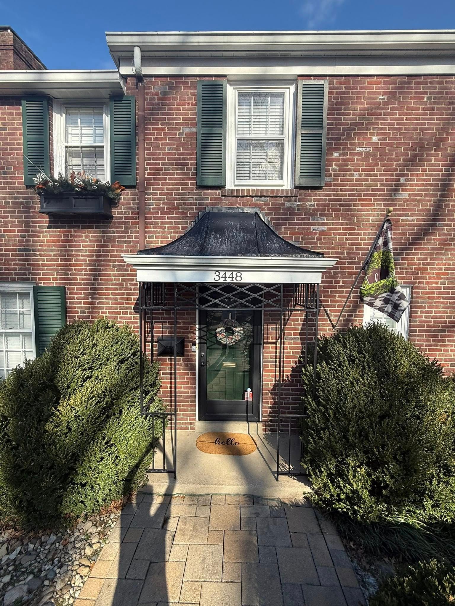 Brick townhouse entrance with white trim, green shutters, and a front walkway bordered by shrubs.