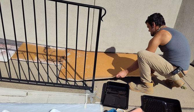 A person in a gray tank top and tan pants kneeling on a patio to place a cardboard sheet near a black metal railing.