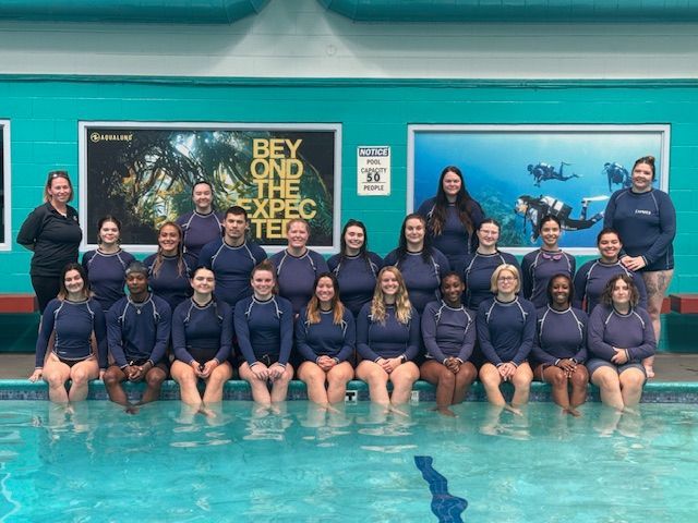 Group of people in a pool wearing blue shirts, posing in front of a turquoise wall with dive-themed posters.