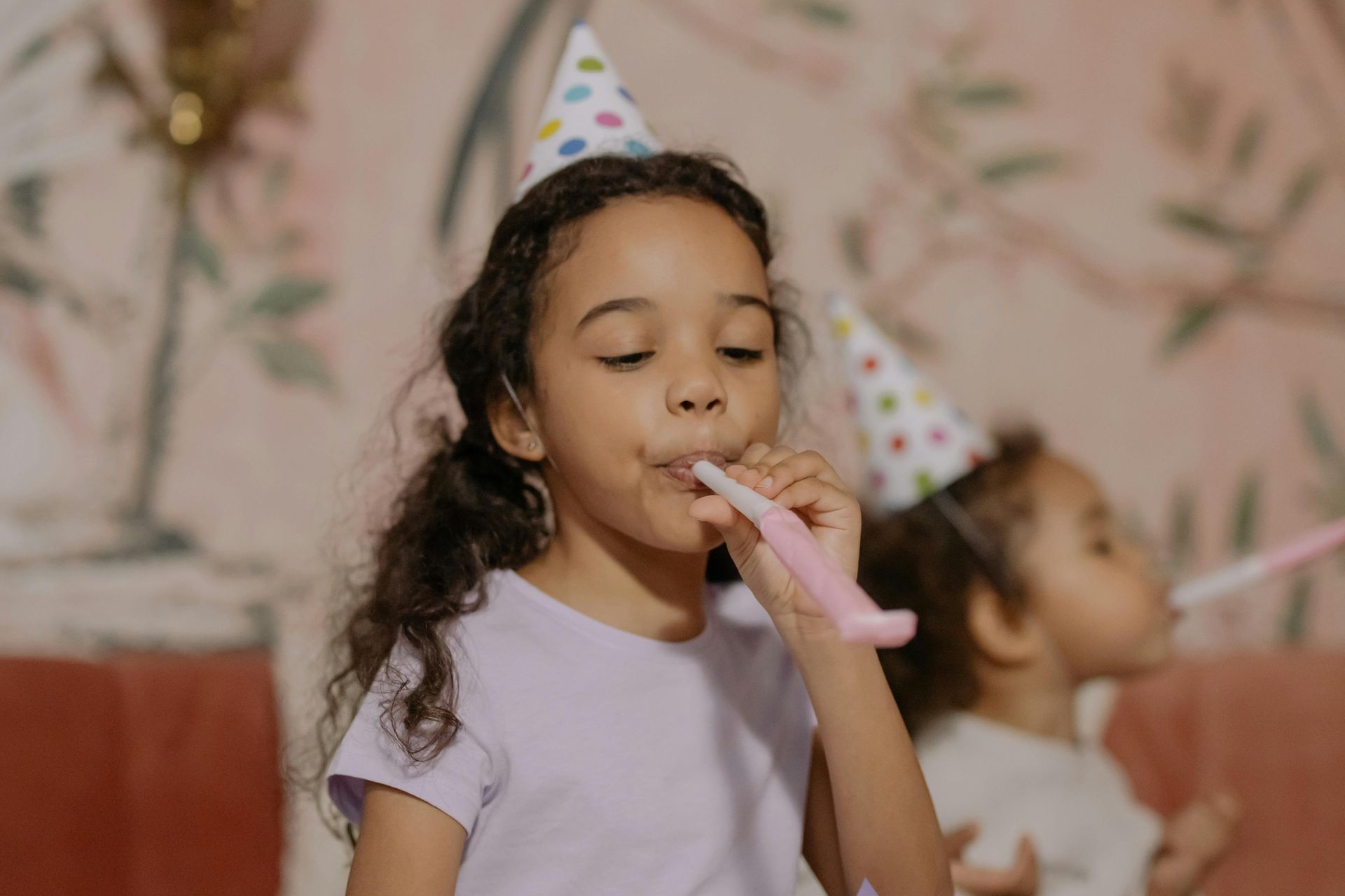 A little girl wearing a party hat is blowing a party horn.