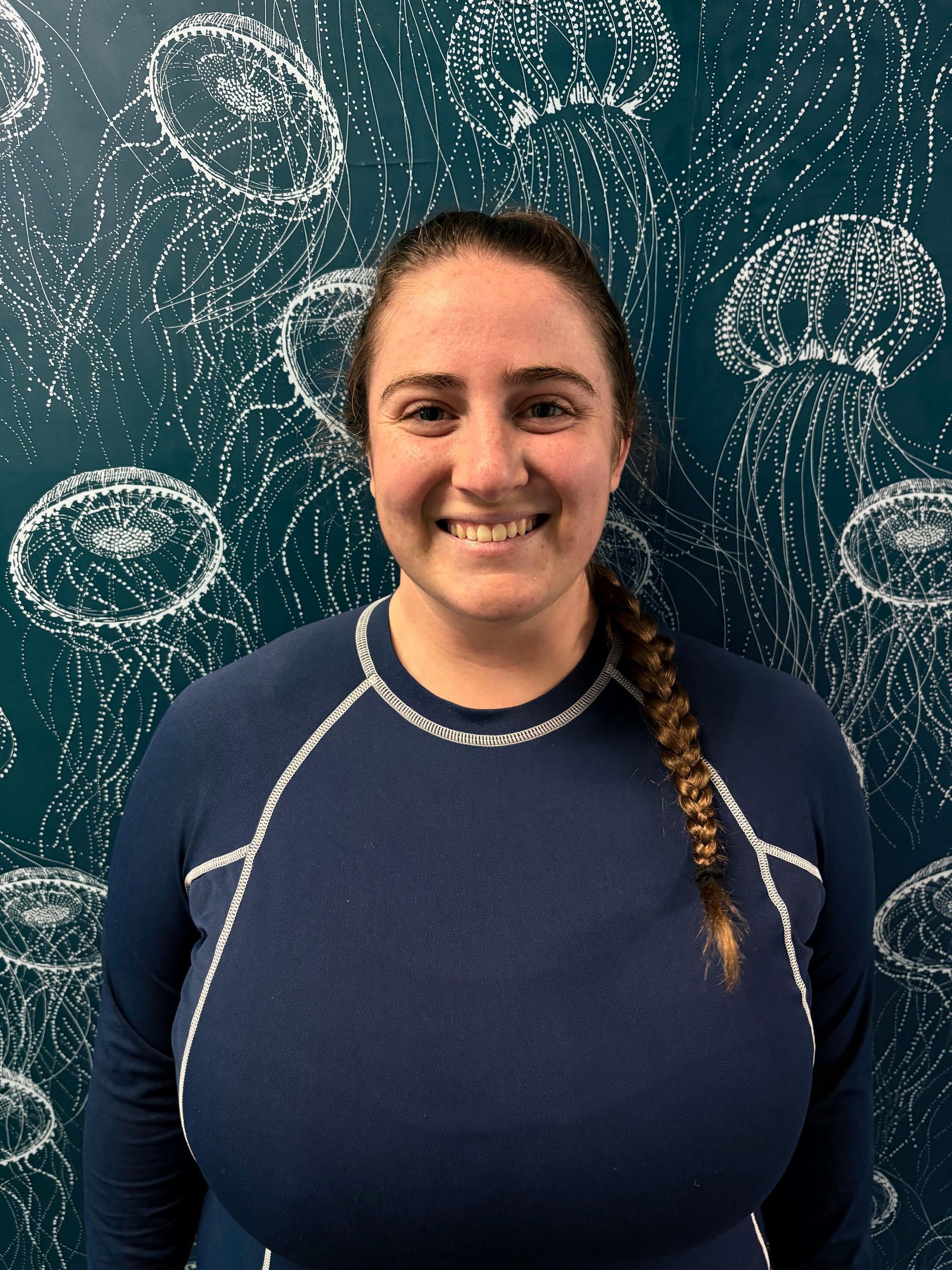 A woman in a blue shirt is smiling in front of a wall with jellyfish on it.