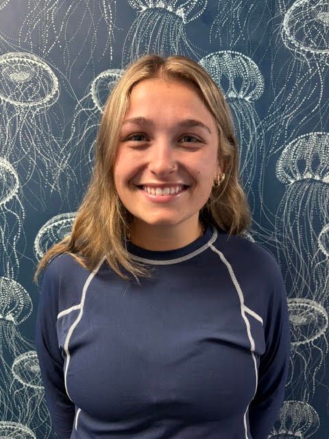 A woman in a blue shirt is smiling in front of a wall with jellyfish on it.