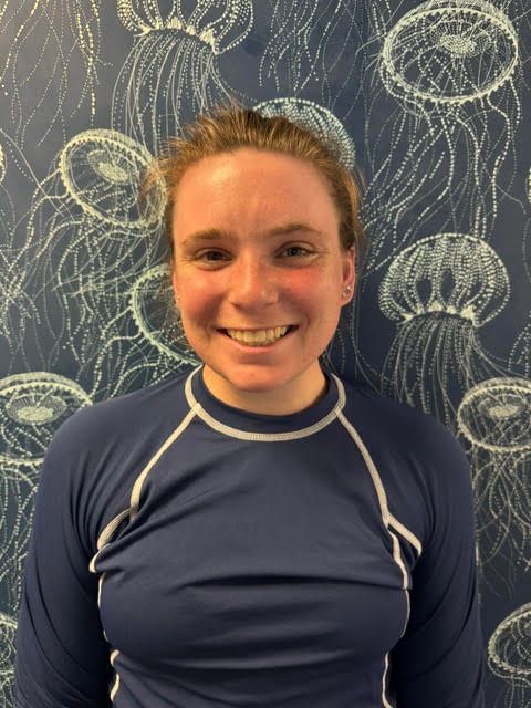 A woman wearing a blue shirt and a necklace is smiling in front of a jellyfish wallpaper.
