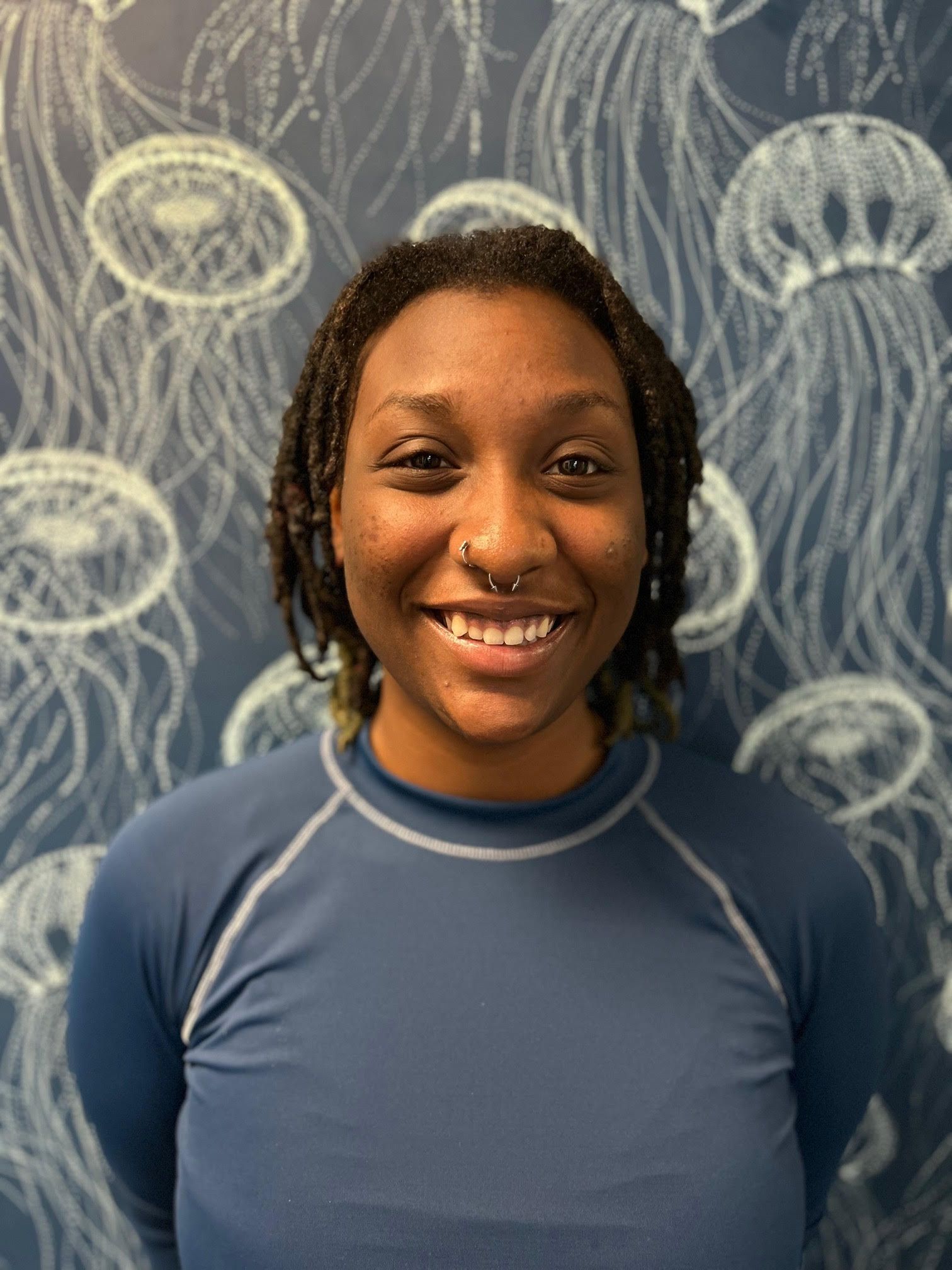 A woman in a blue shirt is smiling in front of a wall with jellyfish on it