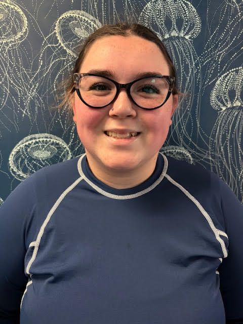 A woman in a blue shirt is smiling in front of a wall with jellyfish on it.
