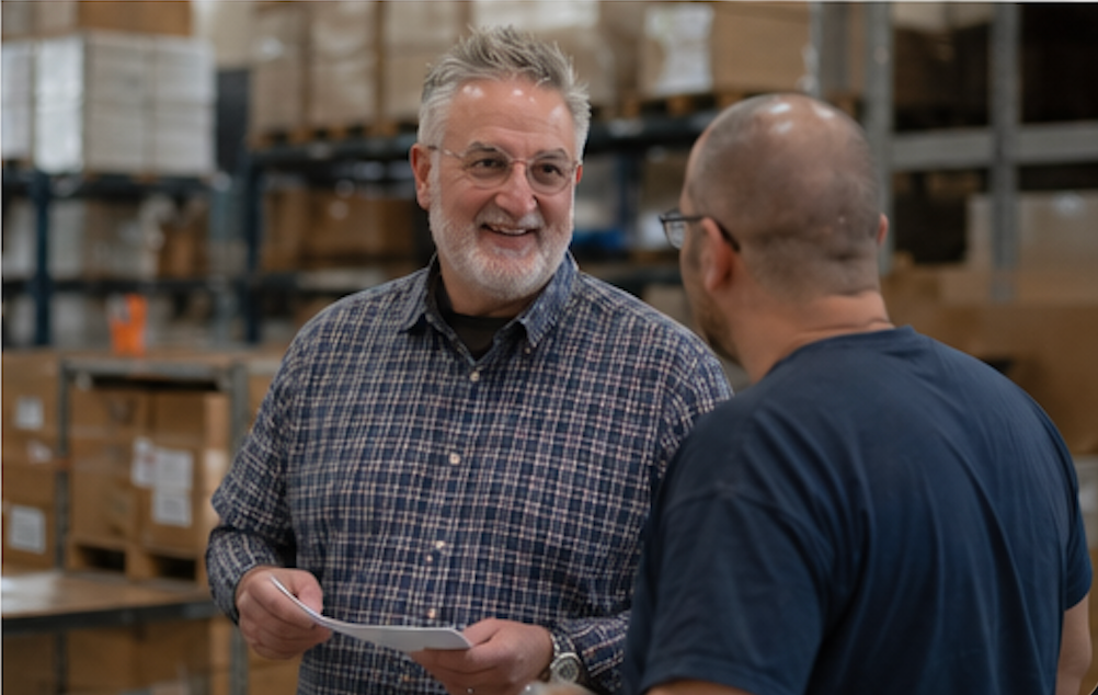 Two men talking in a warehouse, one holding a paper and smiling in front of stacked boxes.