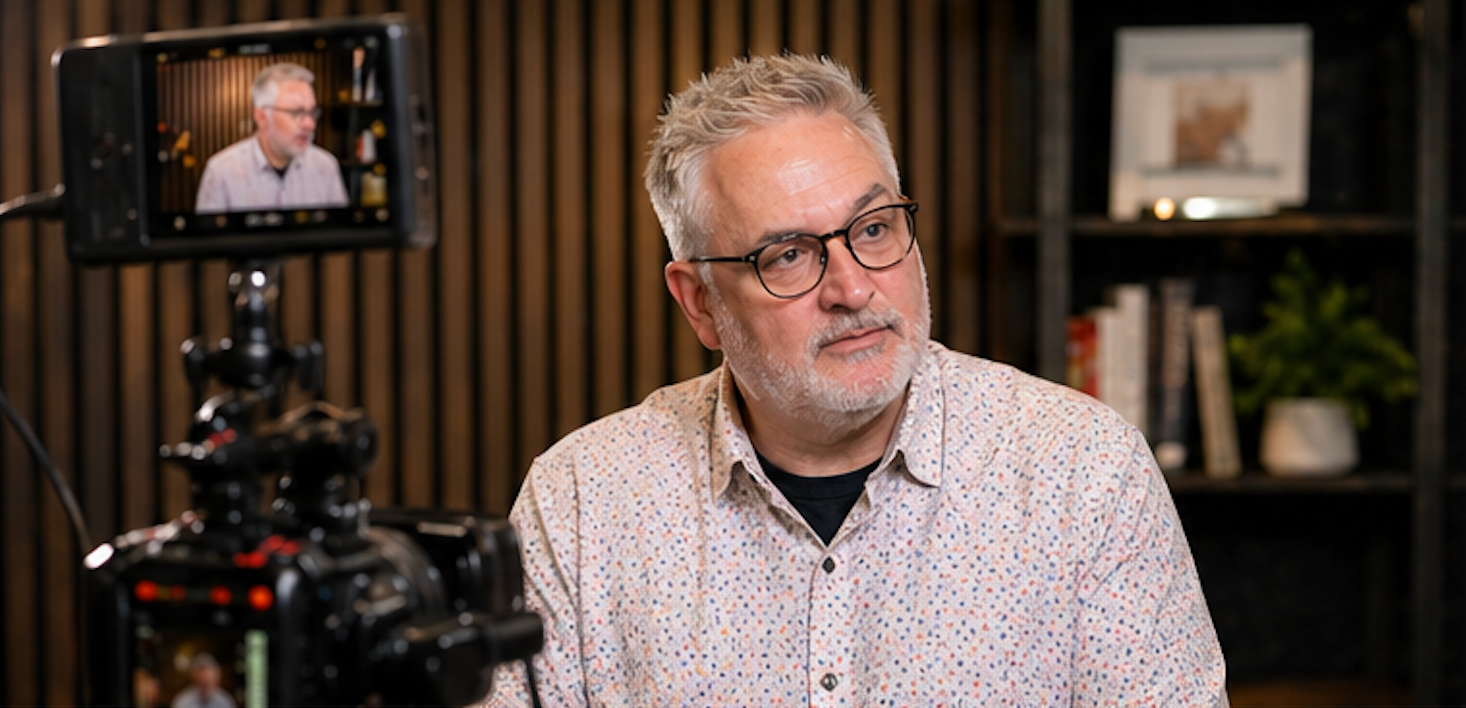 Man in glasses speaking on camera in a studio, with a monitor and camera equipment in the foreground