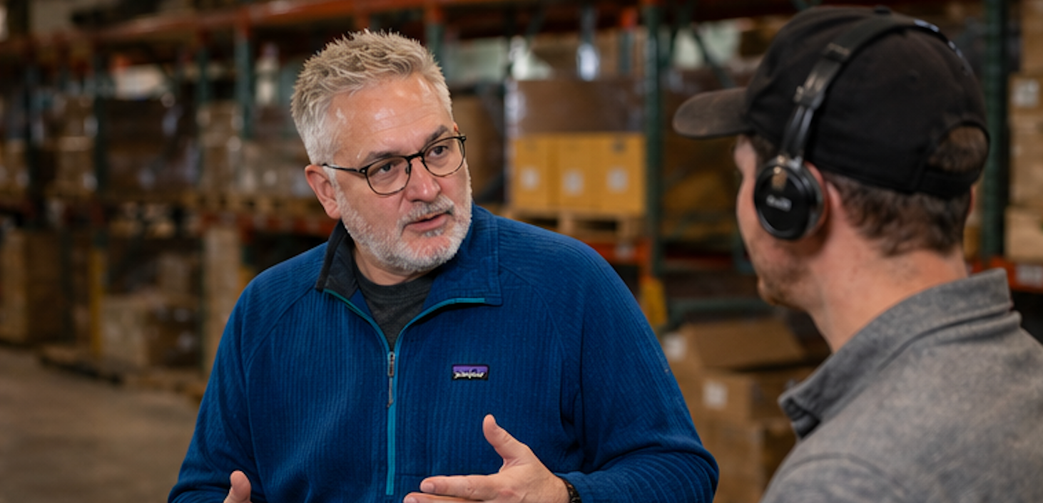 Two people talking in a warehouse, one in a blue jacket gesturing while the other wears headphones.
