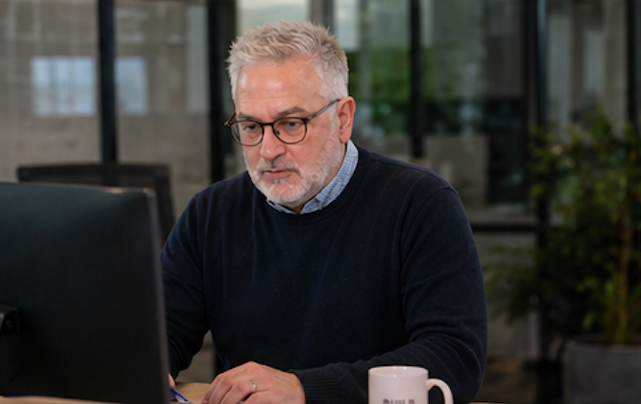 Man working at a desk on a laptop, wearing glasses and a dark sweater in an office setting