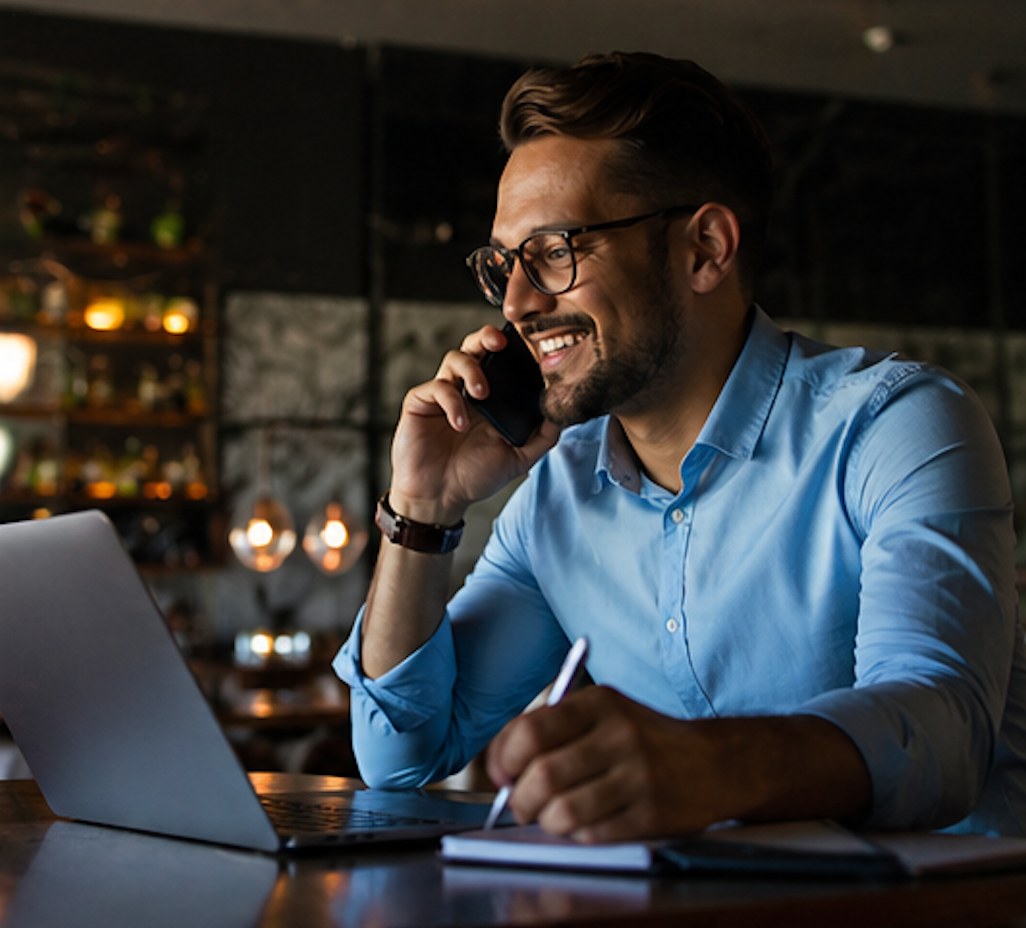 Man in a blue shirt smiling on a phone call while working on a laptop in a dimly lit café