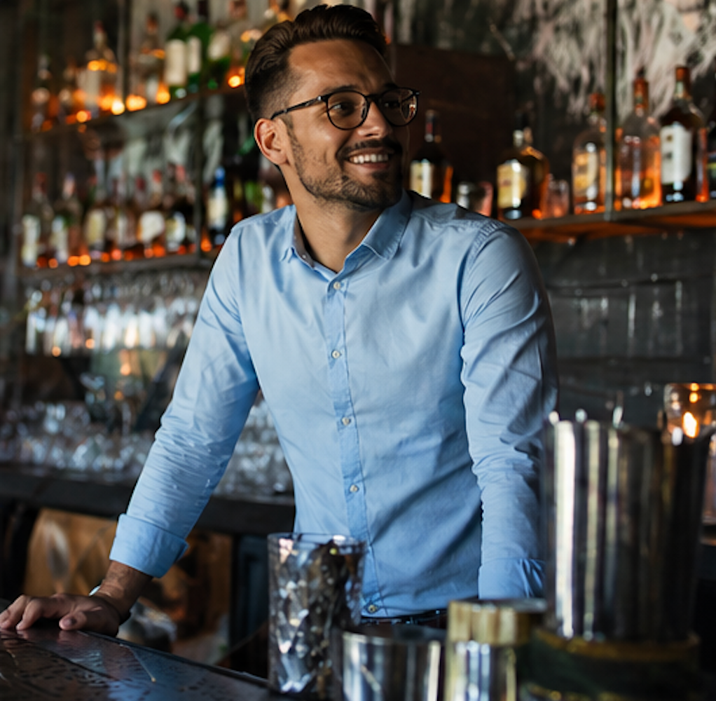 Bartender in a light blue shirt smiling behind a bar with bottles and metal shakers