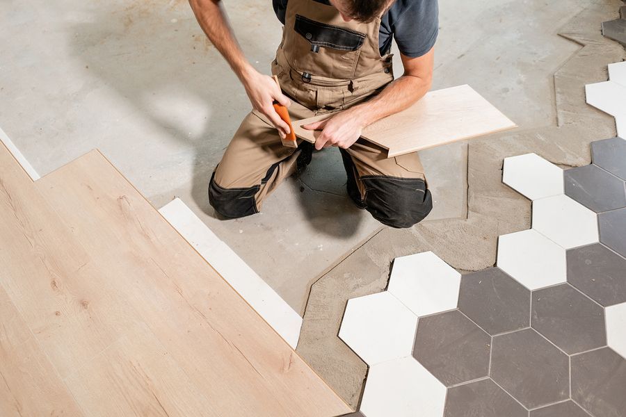 Person kneeling, cutting flooring. Installing light wood planks next to hexagon tiles, beige overalls in workshop.