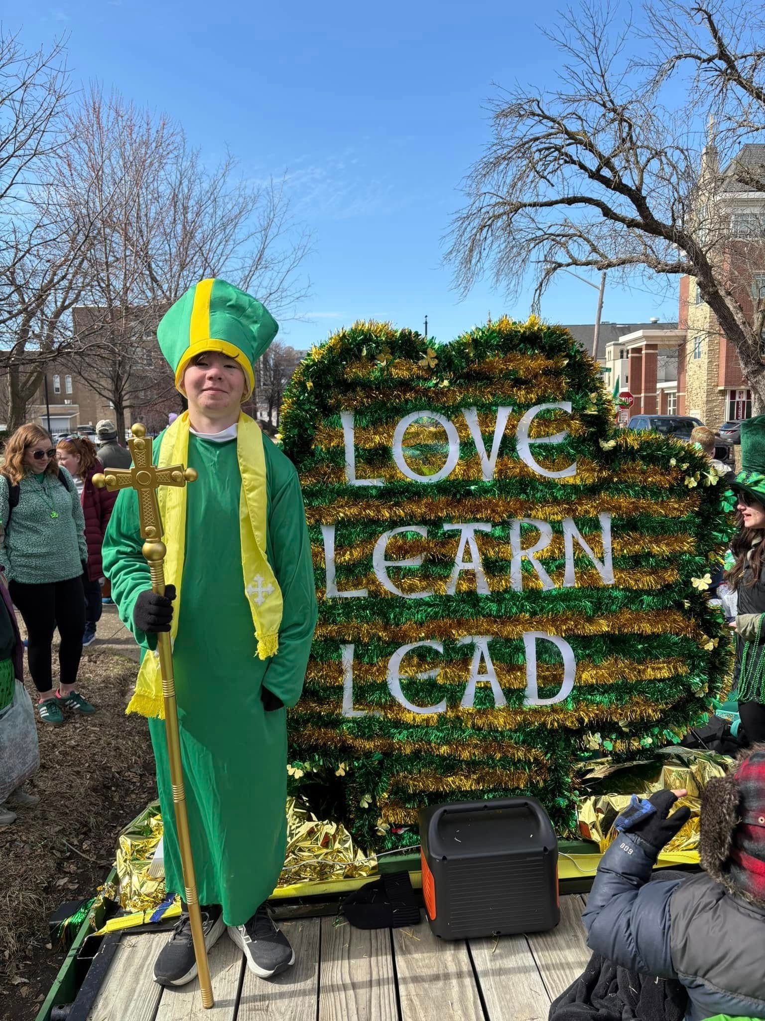 Person in green robe and hat with staff, in front of a float that says