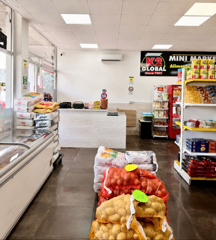 Corridoio di un piccolo supermercato con sacchetti di frutta e verdura in primo piano e scaffali di snack sulla destra.