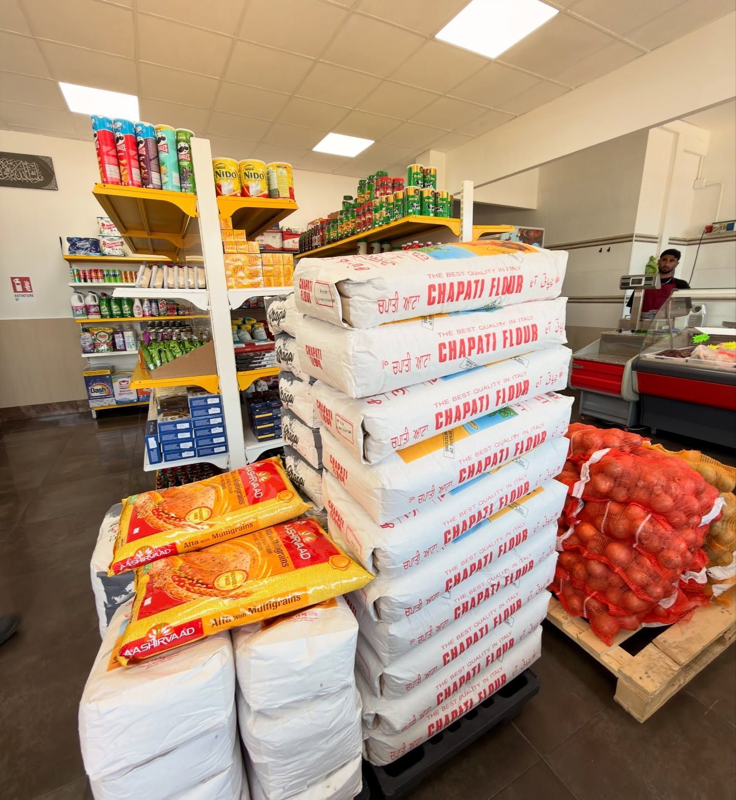 Corridoio di un supermercato con pile di sacchi e scatole di fagioli, riso e prodotti ortofrutticoli.