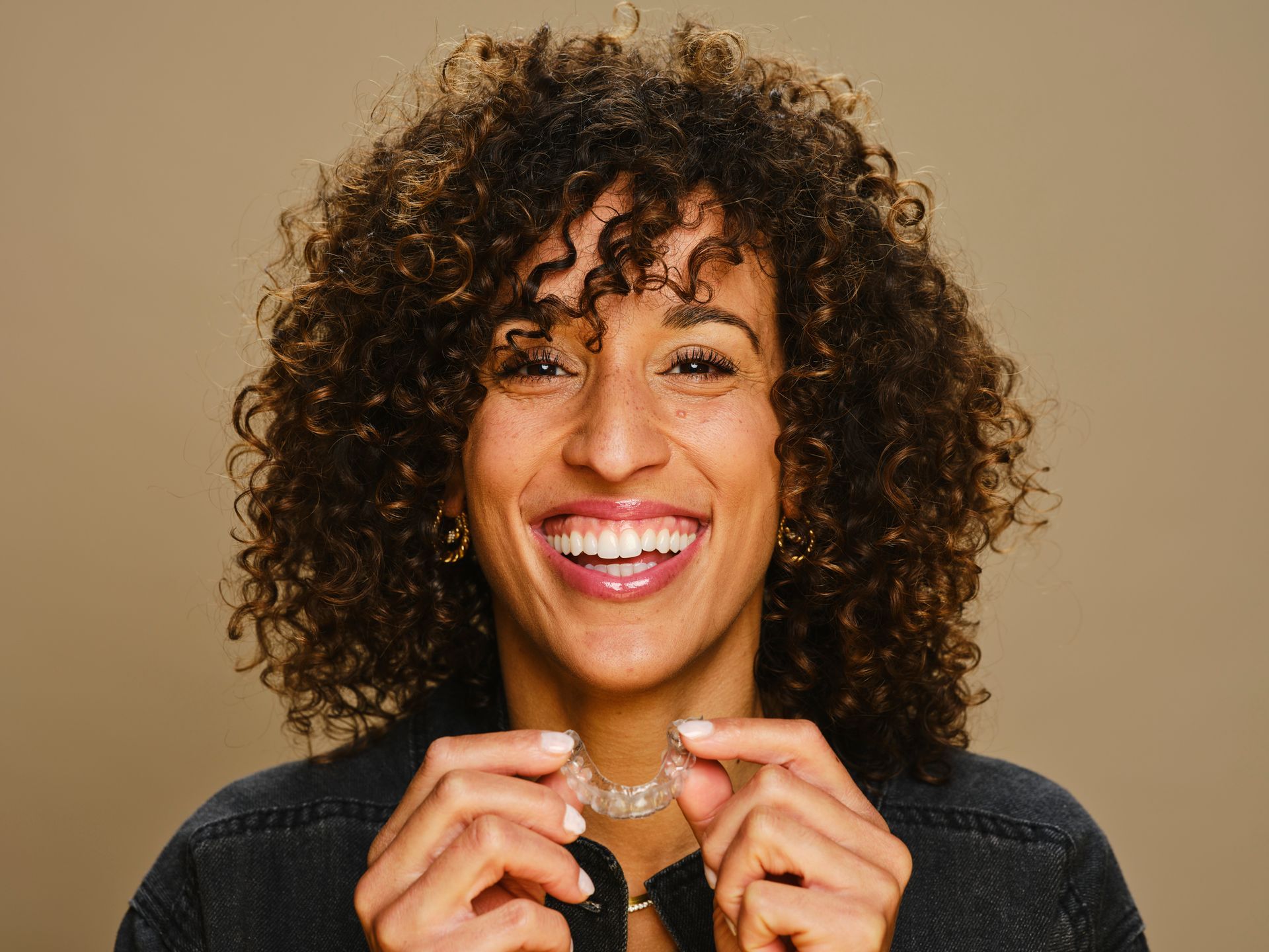 Woman with curly hair smiling, holding clear aligners, beige background.