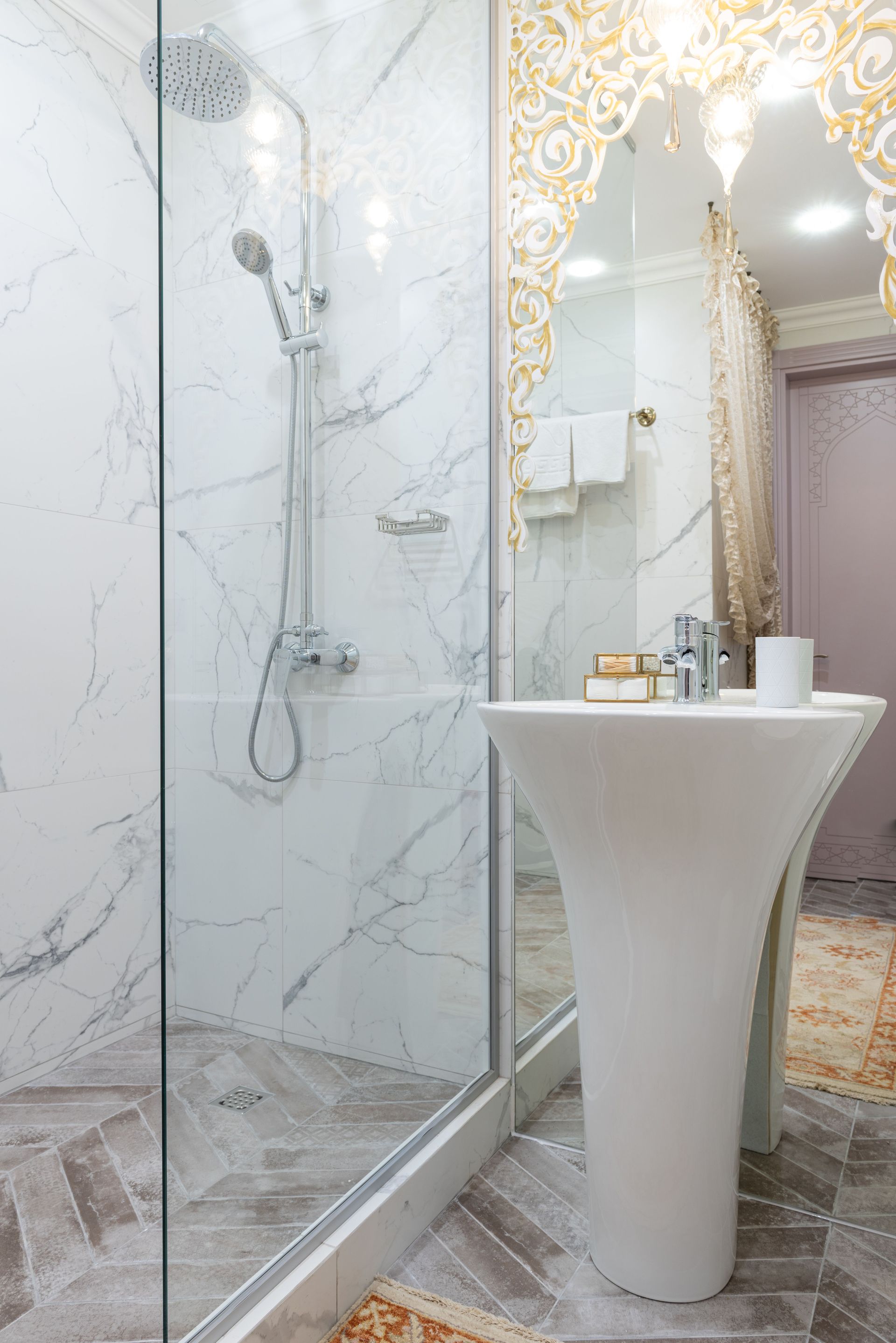 Elegant bathroom with glass shower, marble walls, and a white pedestal sink in a bright interior.