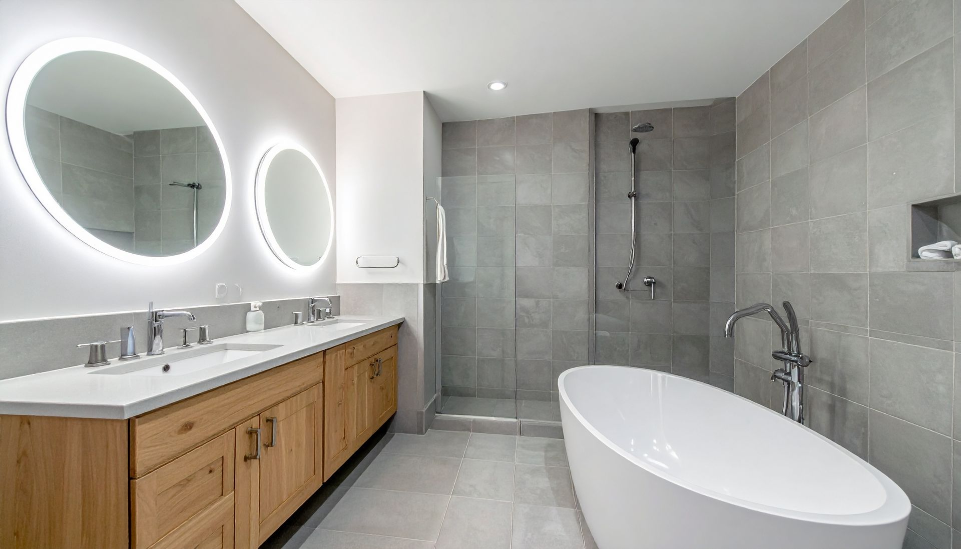 Modern bathroom with double vanity, round lit mirrors, gray tile, and a freestanding tub