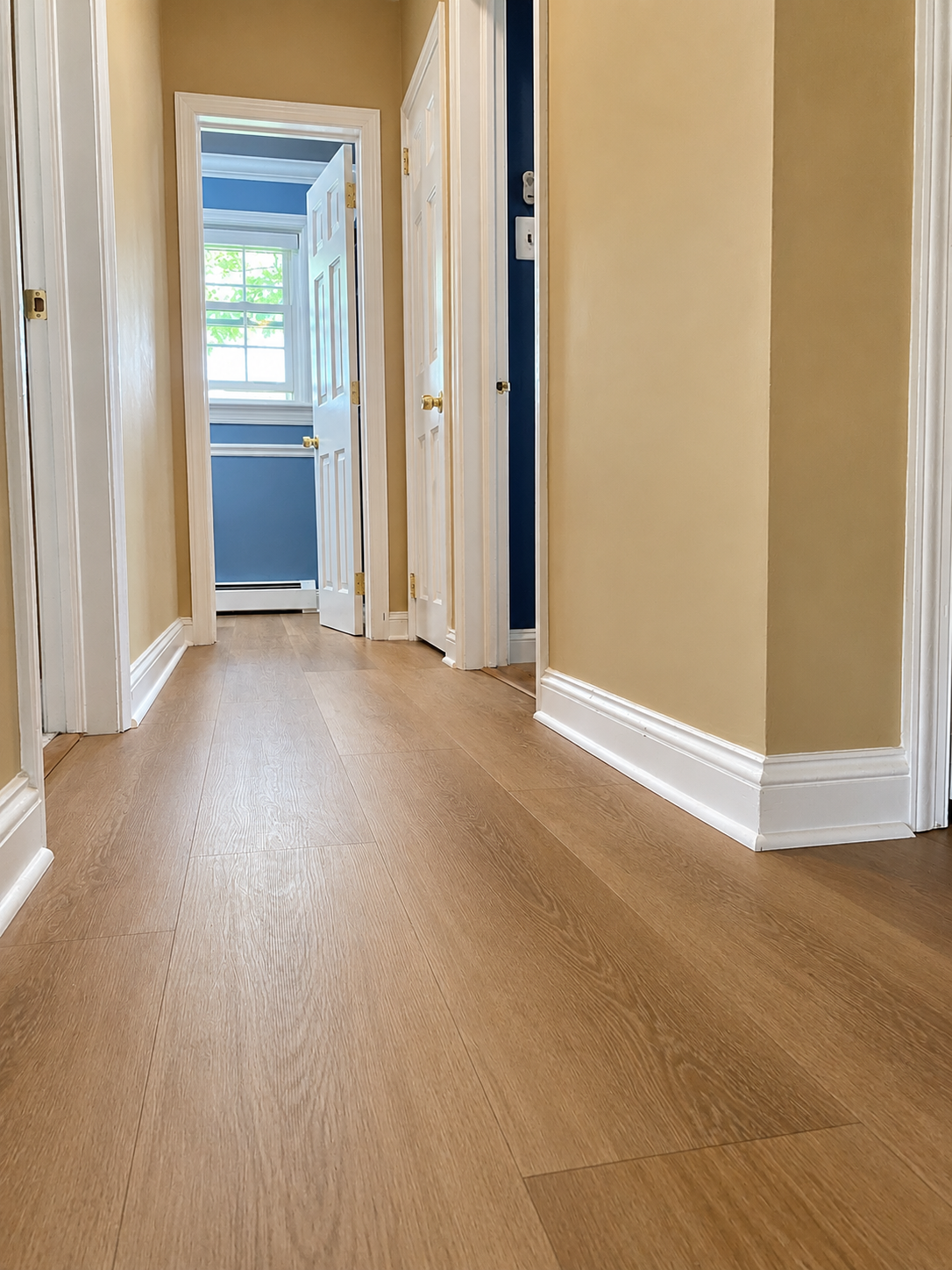 Empty hallway with light wood floors, beige walls, and a view into a bright room at the end.