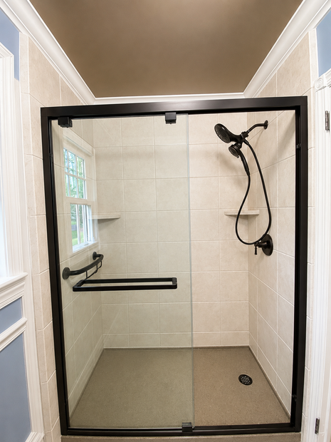 Small tiled walk-in shower with black trim, glass door, grab bars, and handheld showerhead.