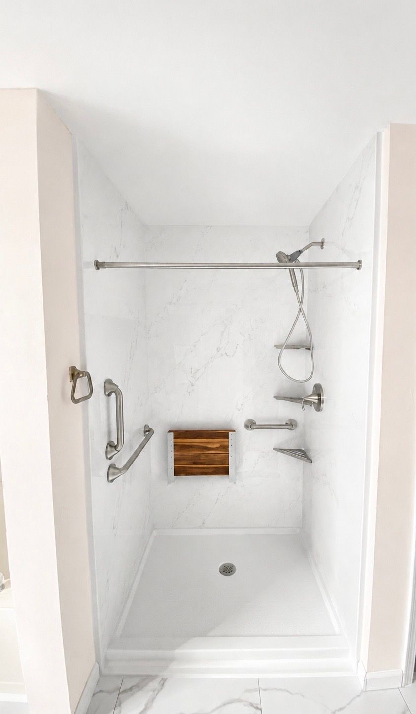 Small white tiled shower stall with grab bars, bench, handheld showerhead, and wooden seat panel.