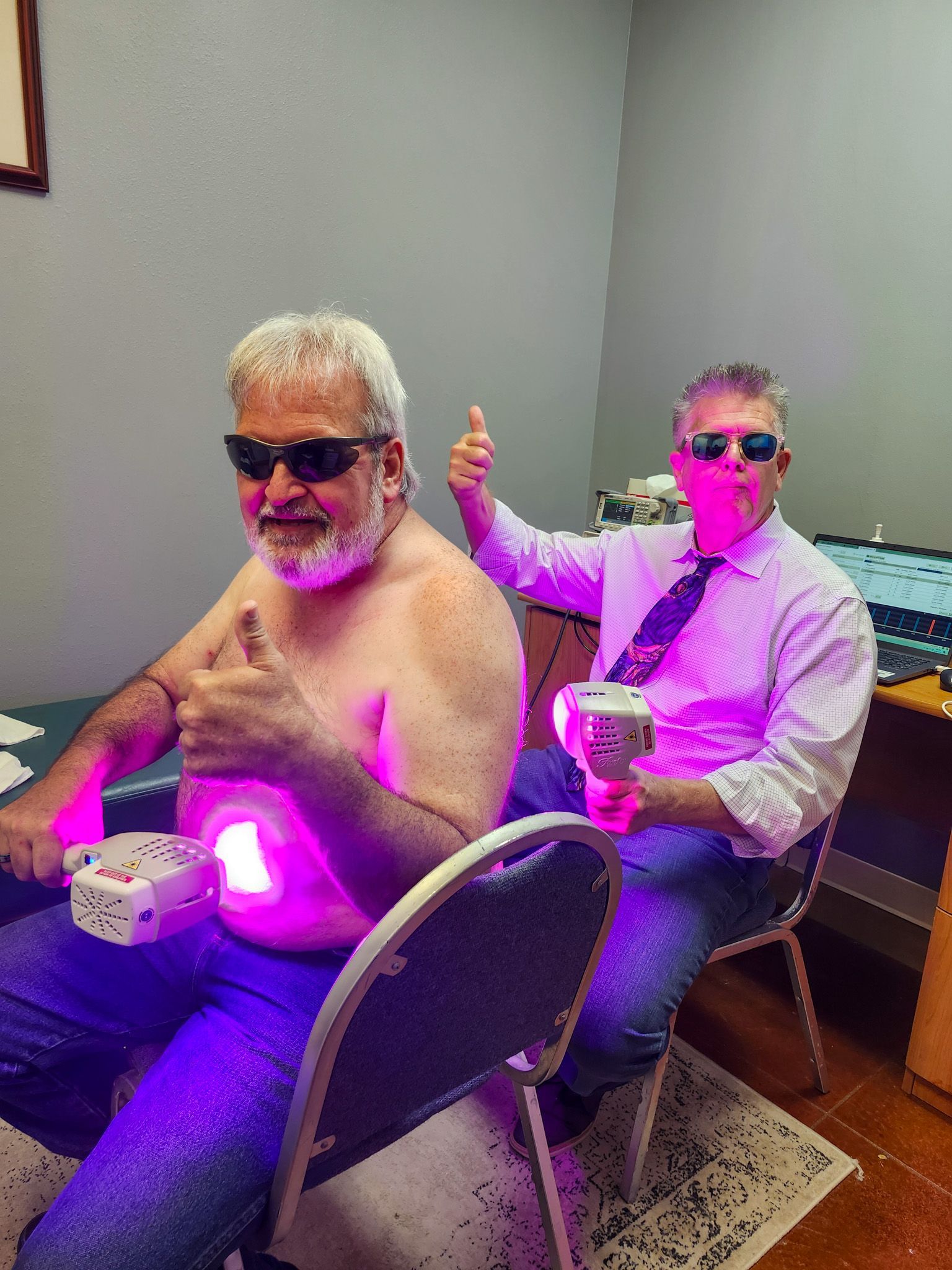 Two men are sitting in chairs in a room with purple lights on their bodies.