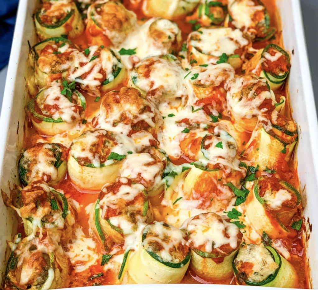 A casserole dish filled with stuffed zucchini rolls covered in sauce and cheese.