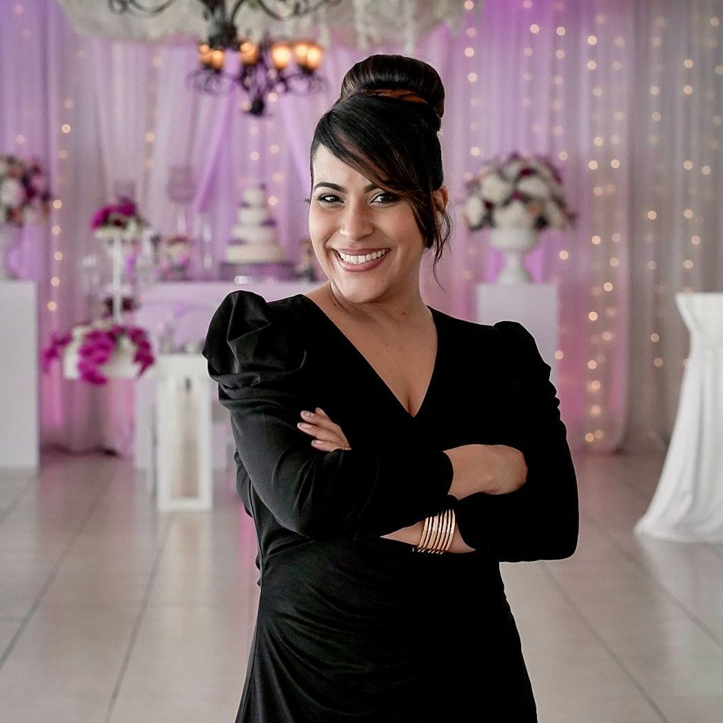 Woman smiling with arms crossed, in a black dress, in a decorated event space with purple and white decor.