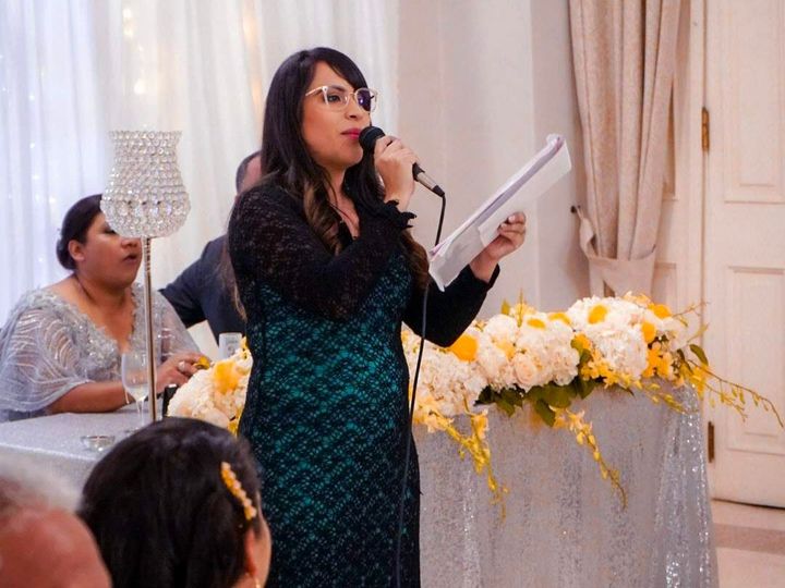 Woman speaking at an event, holding a microphone and papers. She wears a black and teal dress, standing behind a decorated table.