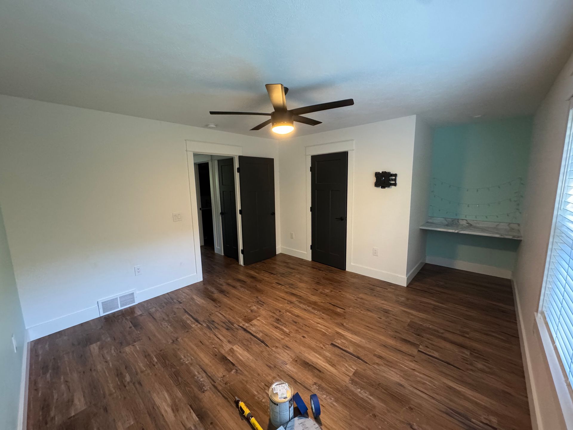 Empty living room with wood floors, ceiling fan, dark doors, and a small built-in desk area.