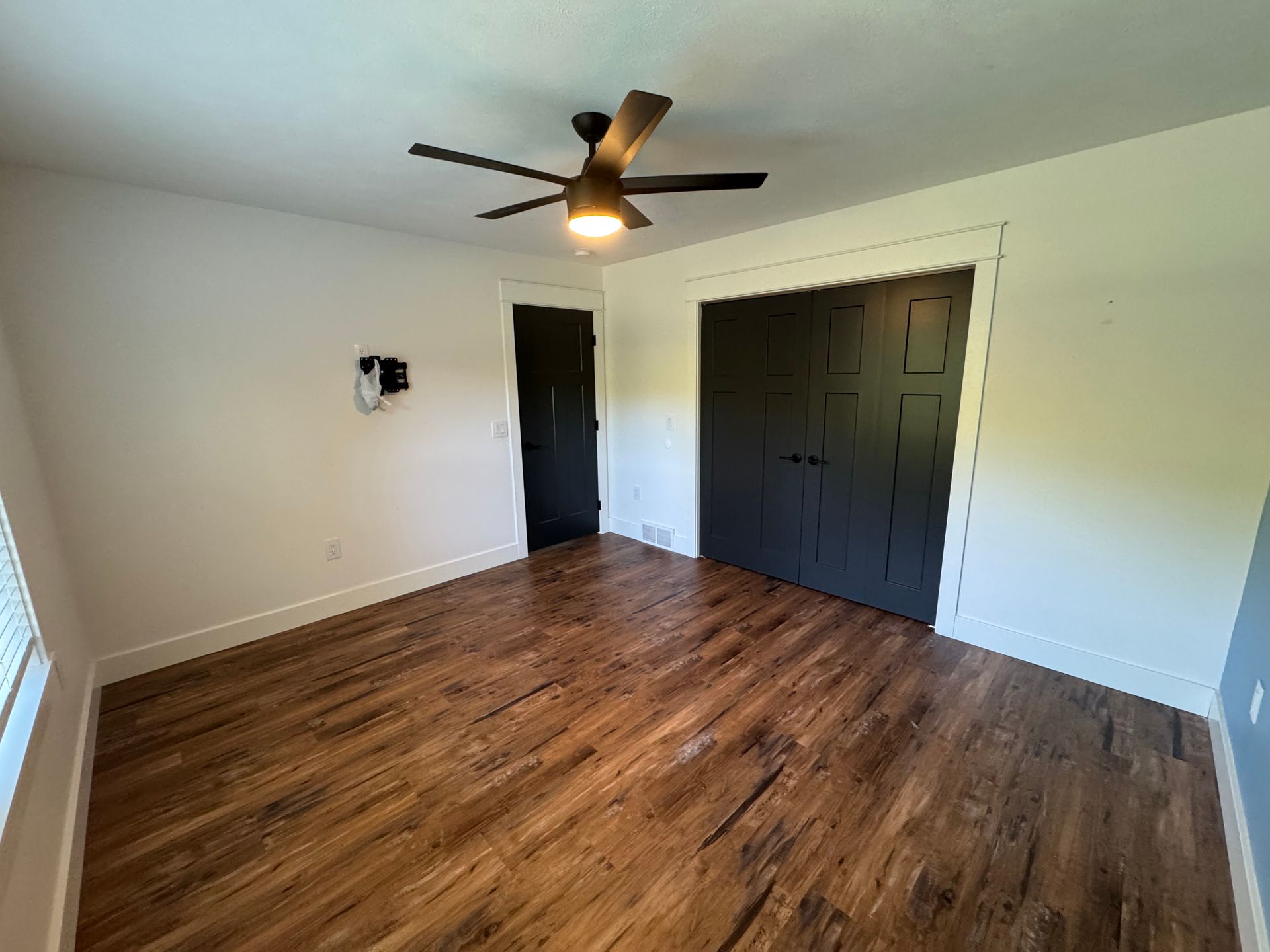 Empty bedroom with wood floors, white walls, ceiling fan, and dark closet doors
