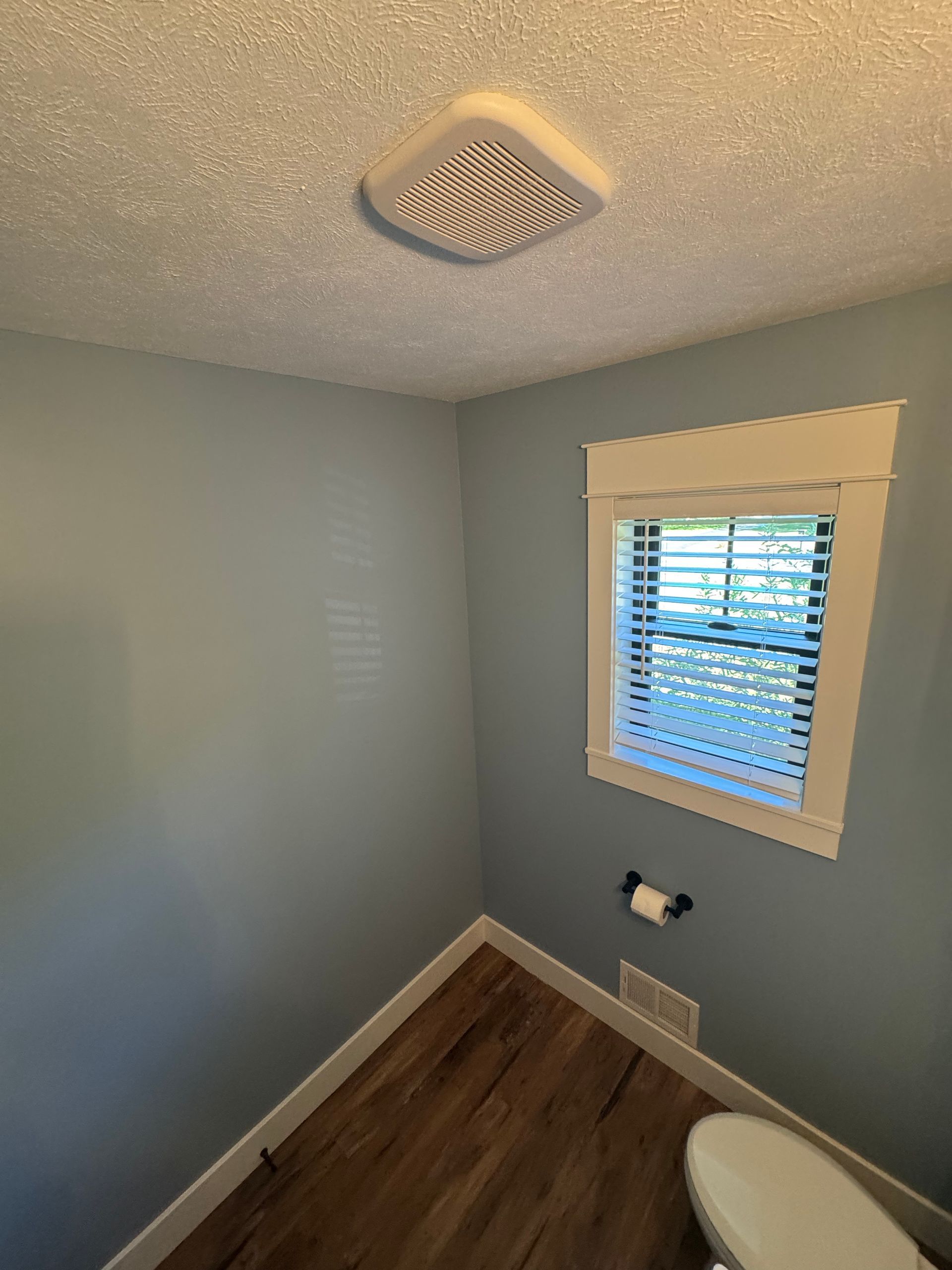 Small bathroom with gray walls, wood floor, window blinds, ceiling vent, and toilet partially visible