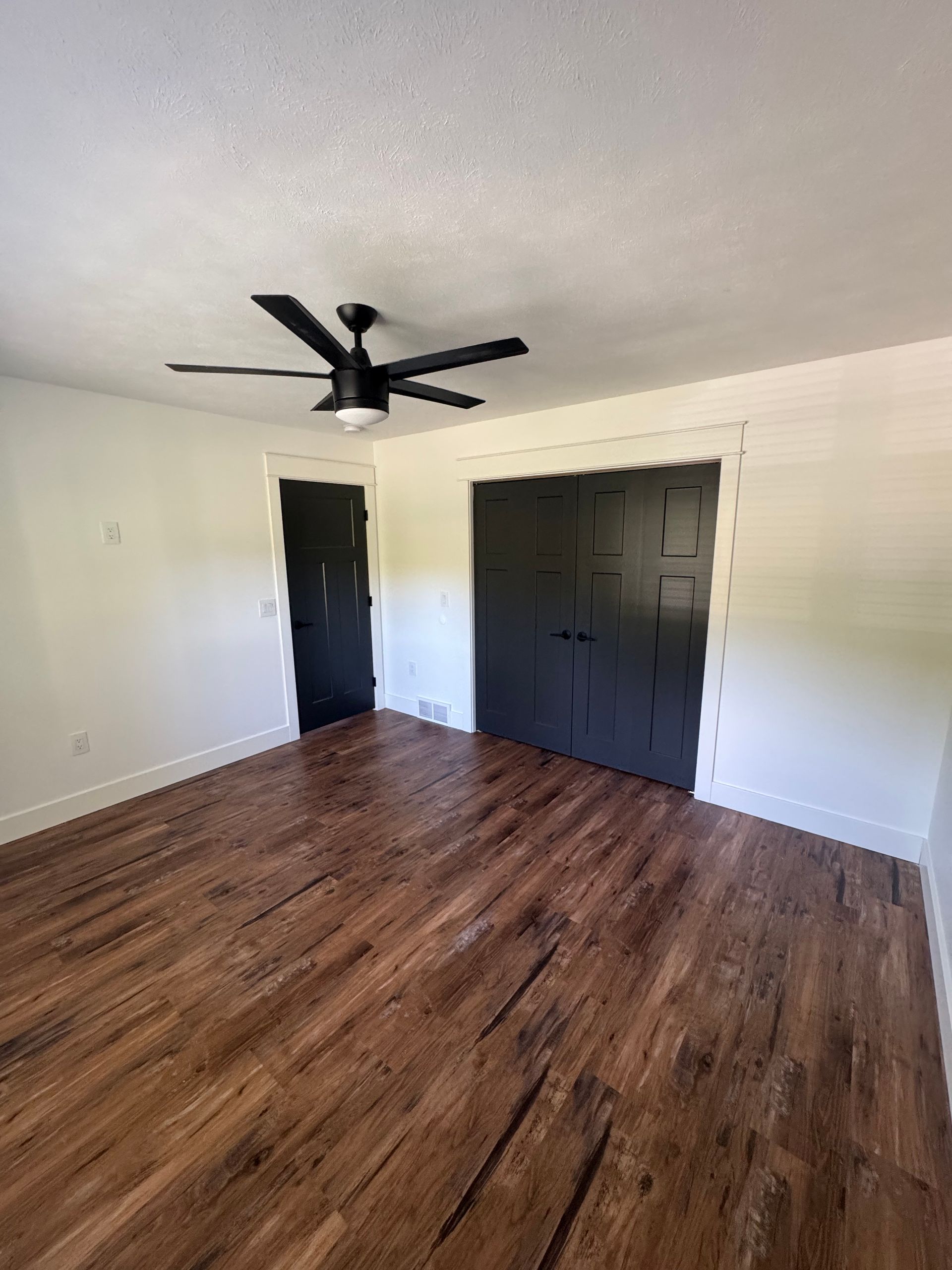 Empty room with wood-look floors, white walls, black ceiling fan, and dark closet doors