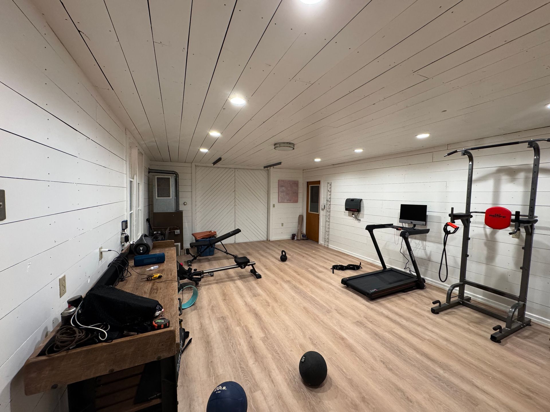 Bright home gym with treadmill, weights, bench, and scattered exercise equipment on wood floor