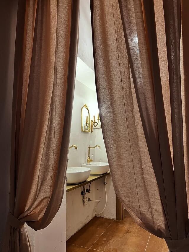 L'ingresso del bagno incorniciato da tende color cuoio. Sono visibili due lavandini bianchi con rubinetti dorati.