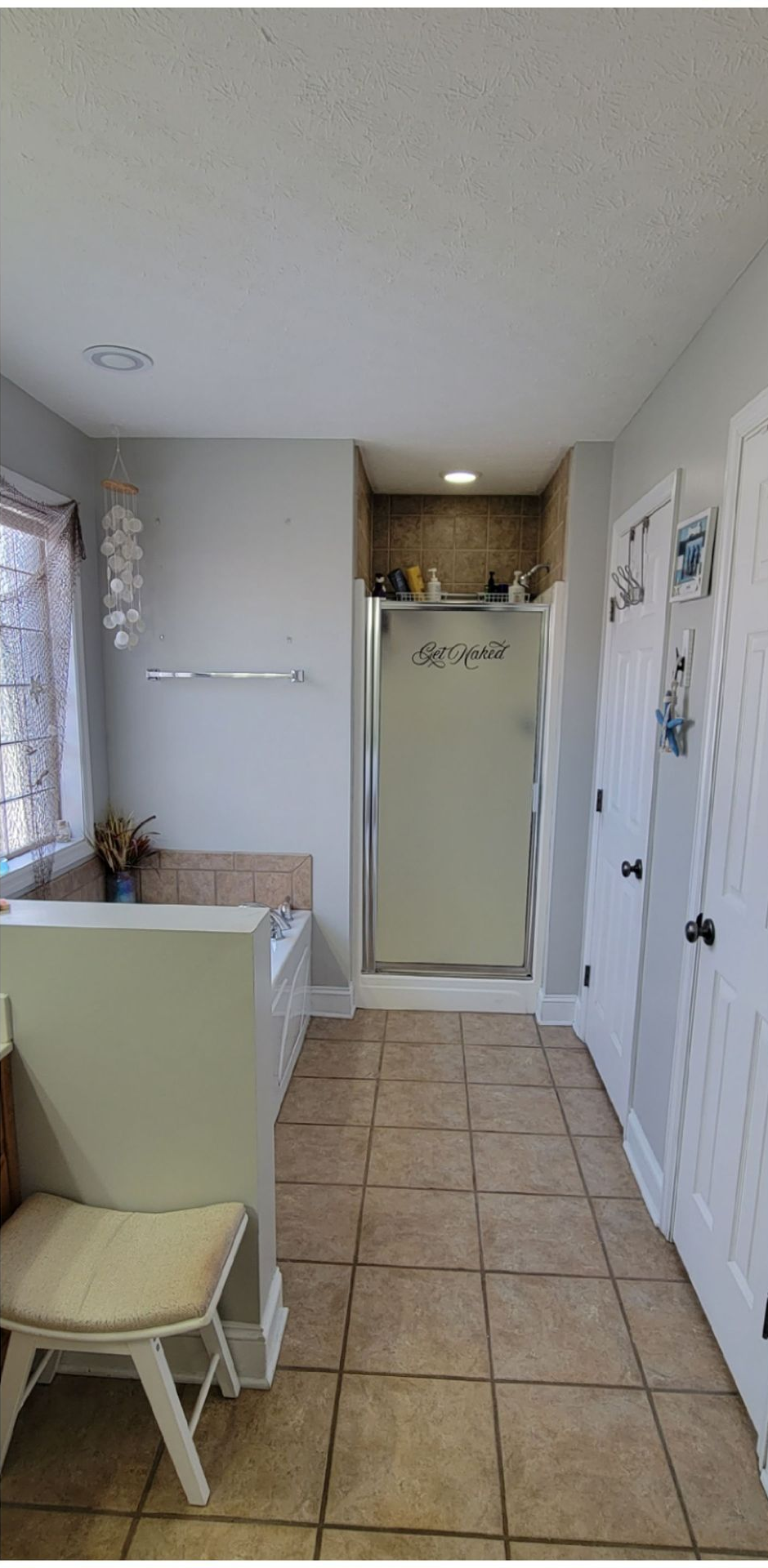 A bathroom with light gray walls, a tan tiled floor, a soaking tub, a glass-enclosed shower, and a small white chair.