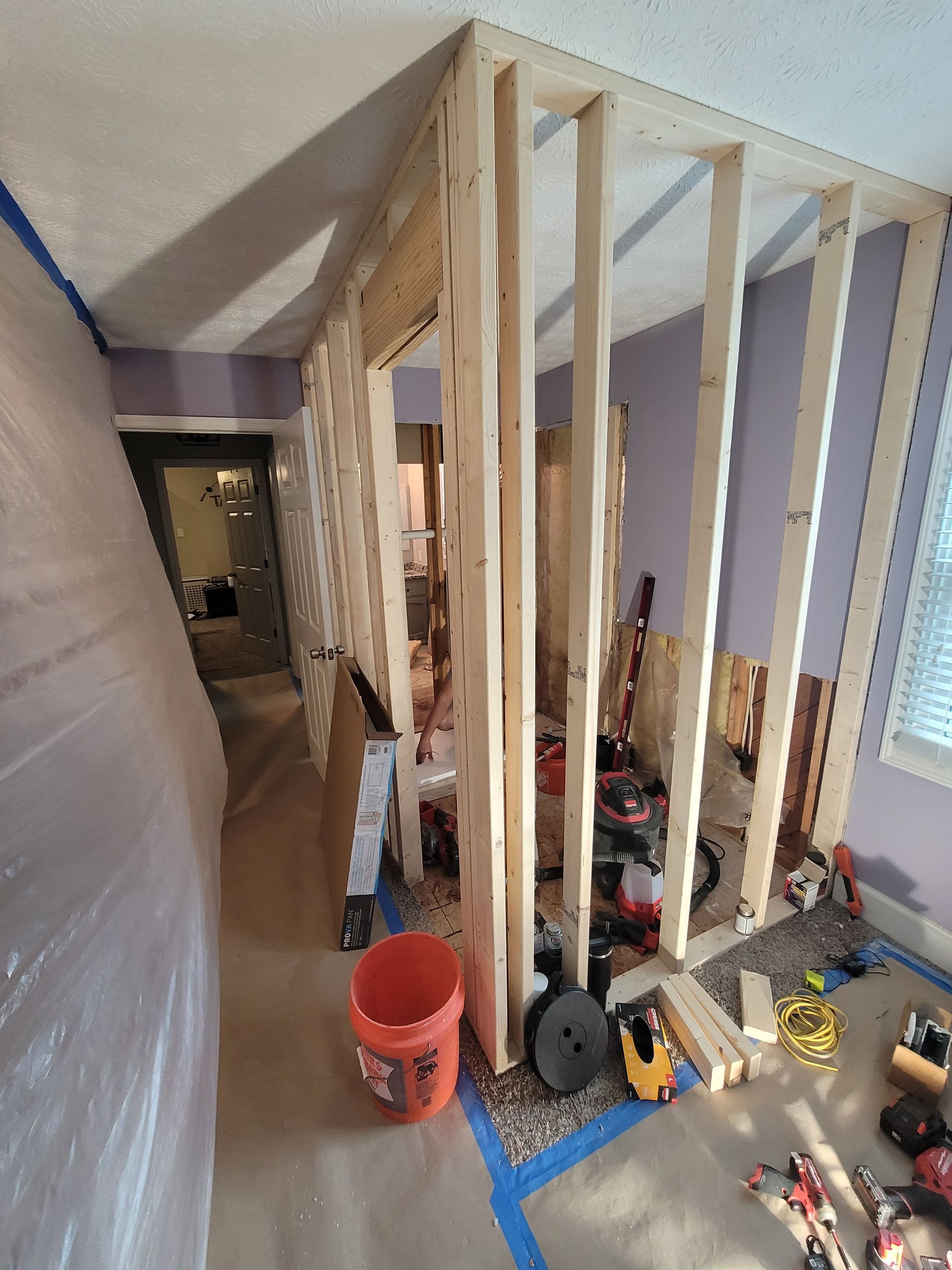 A room undergoing renovation with exposed wooden wall studs framing a doorway, plastic sheeting, and tools on the floor.