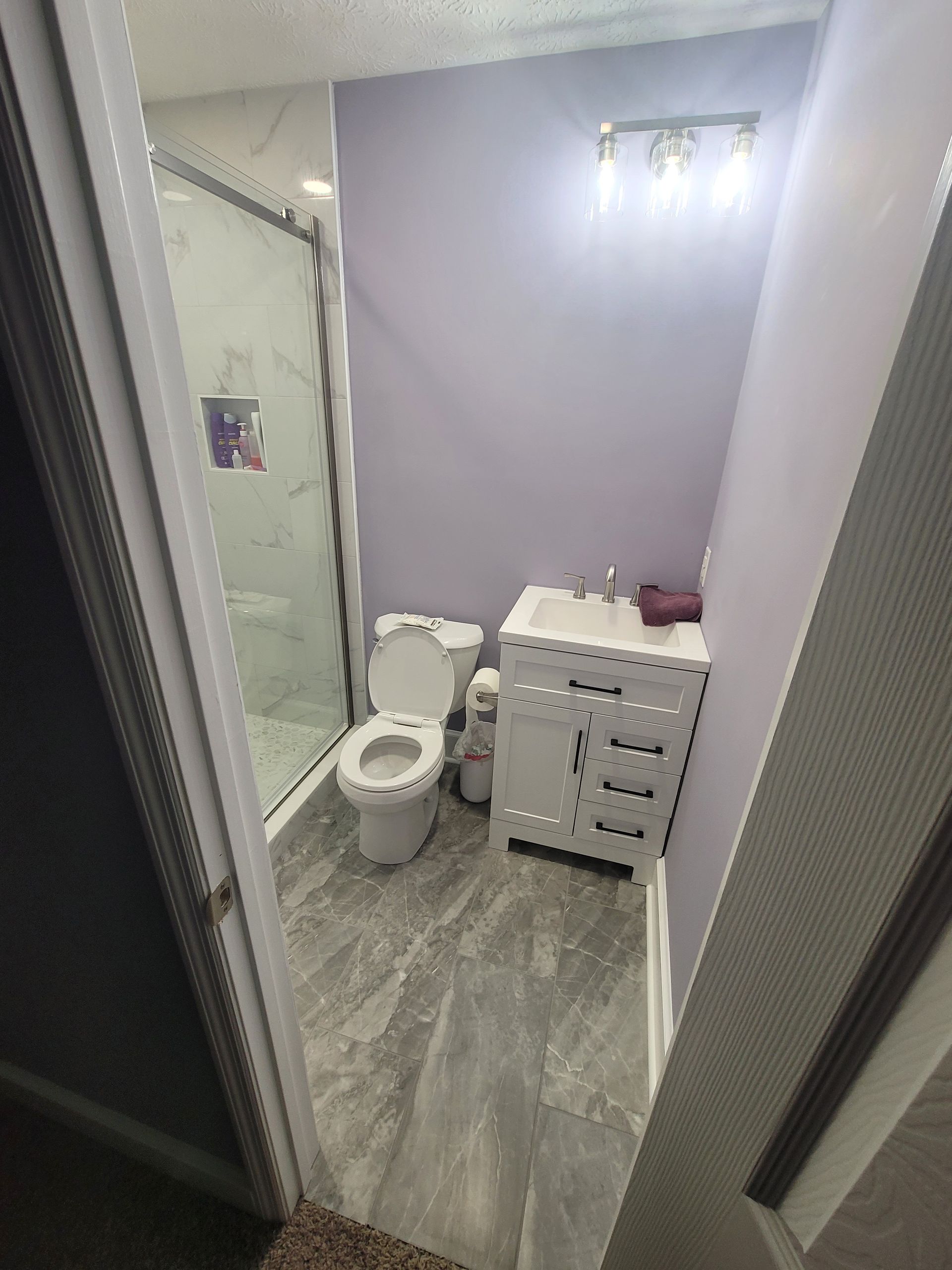 A bathroom featuring grey wood-look flooring, a white vanity, a toilet, and a glass-enclosed shower against a lavender wall.