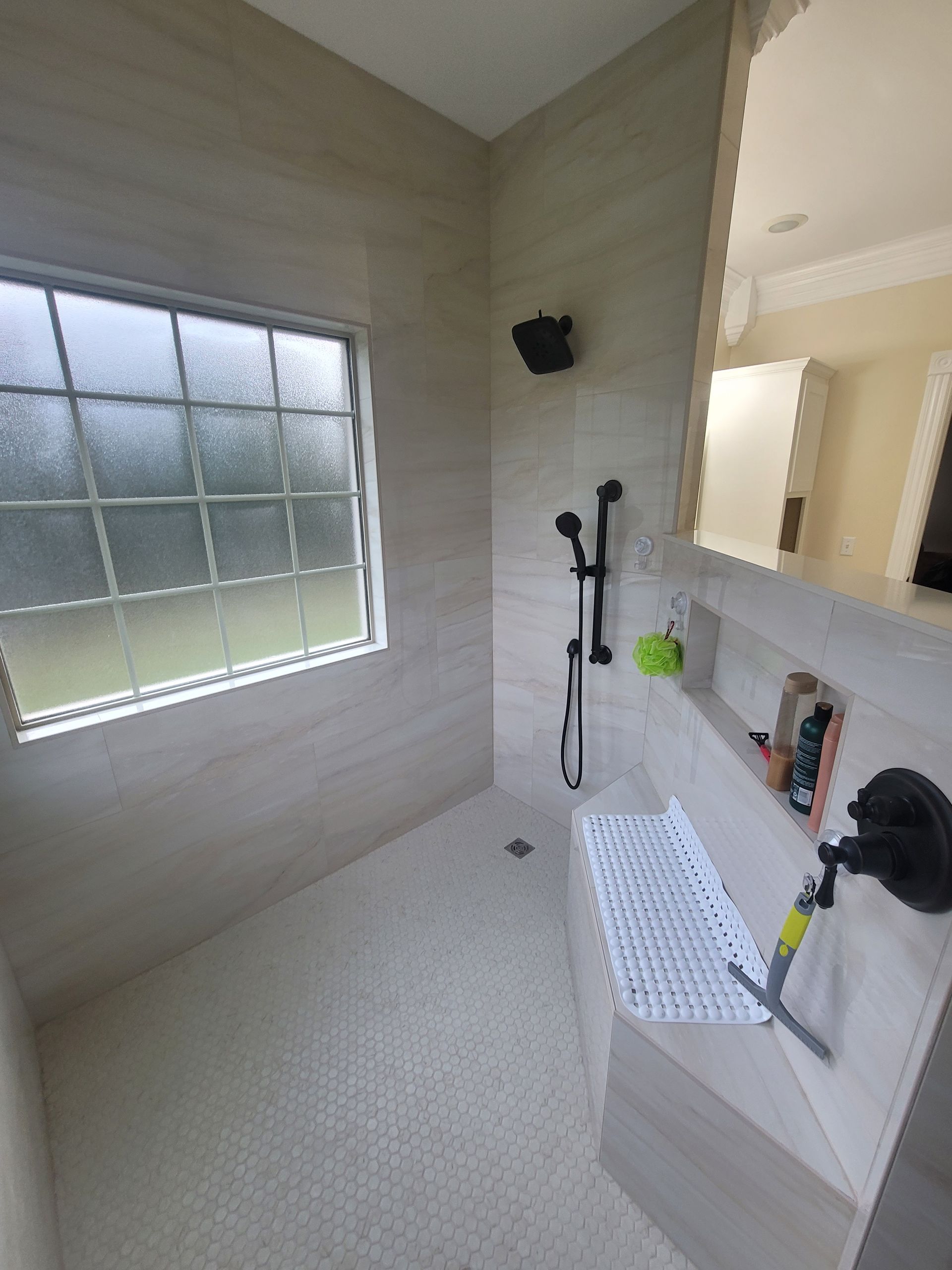 A walk-in shower with light tan stone-look walls, a tiled floor, a handheld showerhead, a built-in bench, and a window.