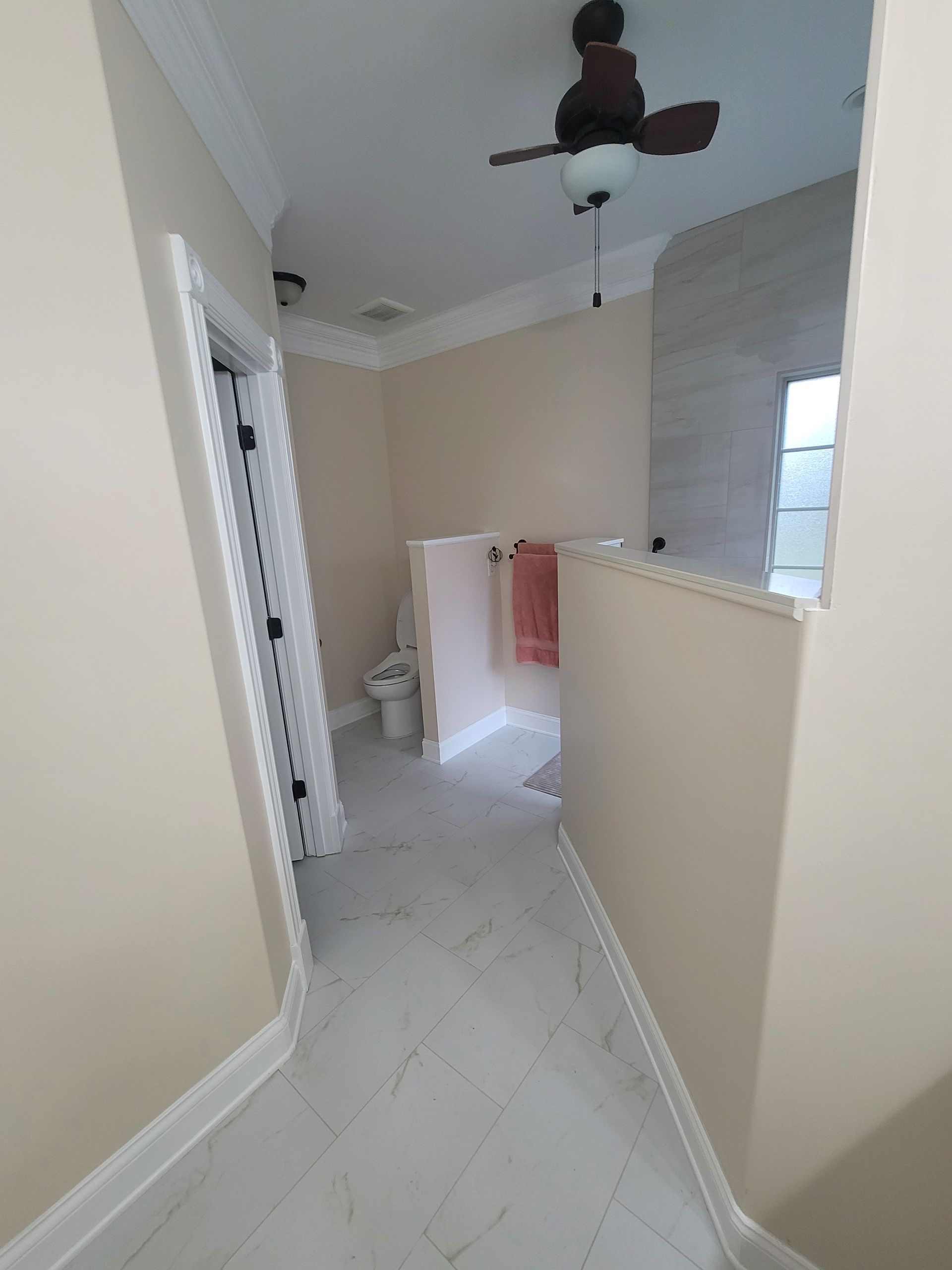 A hallway leads to a bathroom with light tile floors, a toilet, a half-wall, and a ceiling fan.