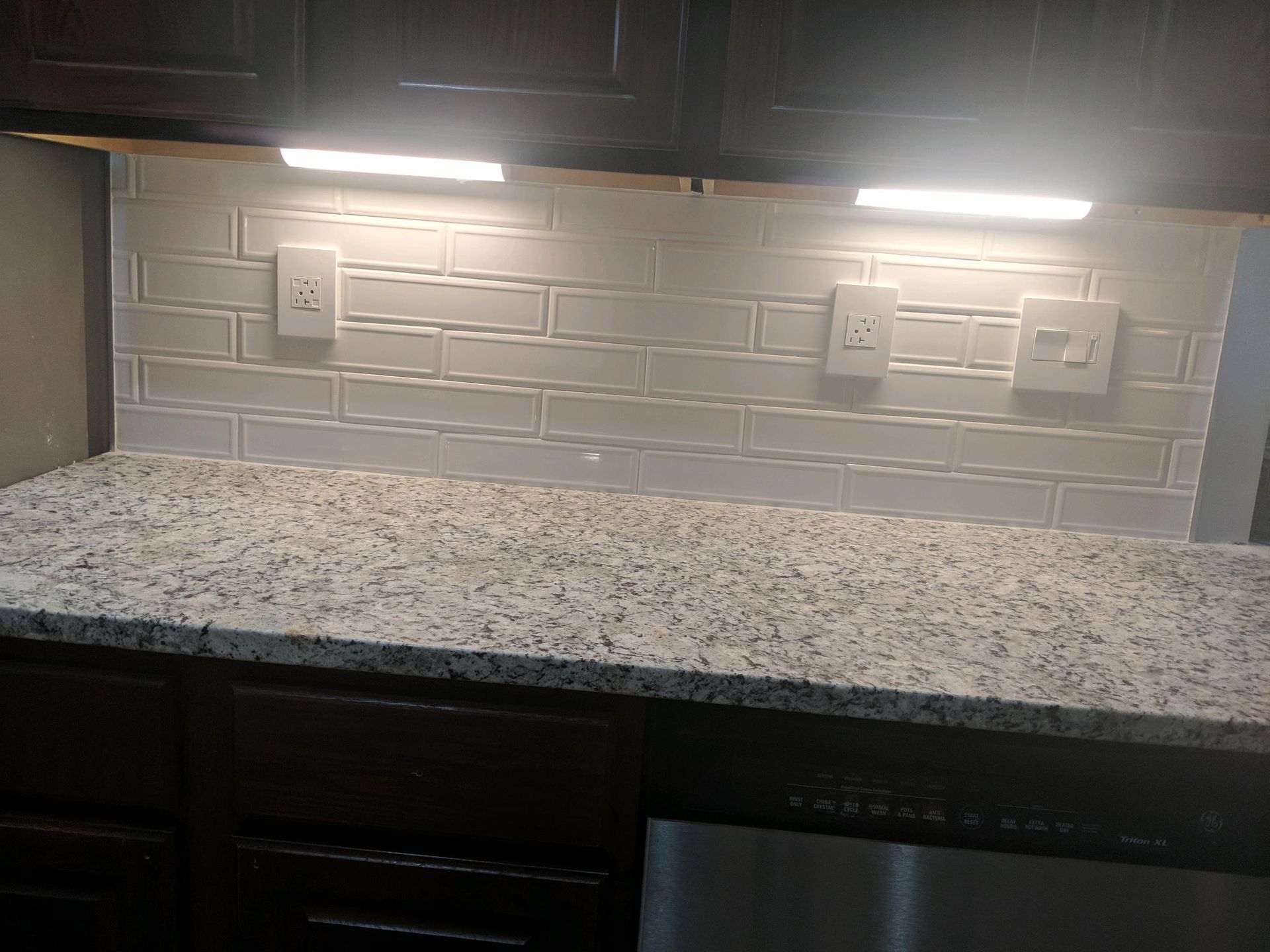 A kitchen counter with light granite, white subway tile backsplash, dark wood cabinets, and three electrical outlets.