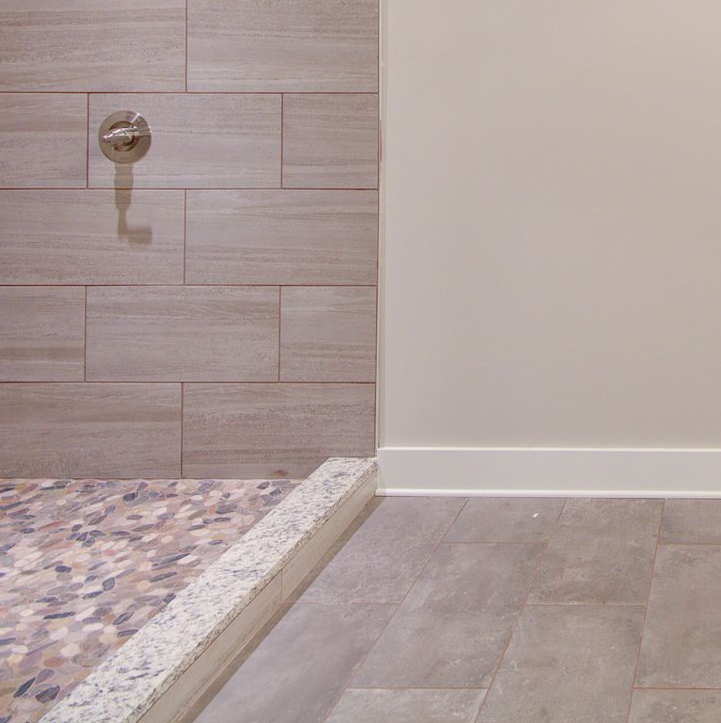 A shower interior with wood-look tile walls and a pebble floor, separated from a grey-tiled bathroom floor by a stone curb.
