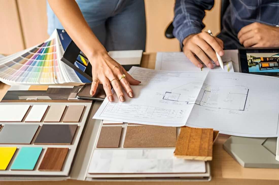 Two people reviewing interior design plans and material samples on a desk filled with color swatches and wood finishes.