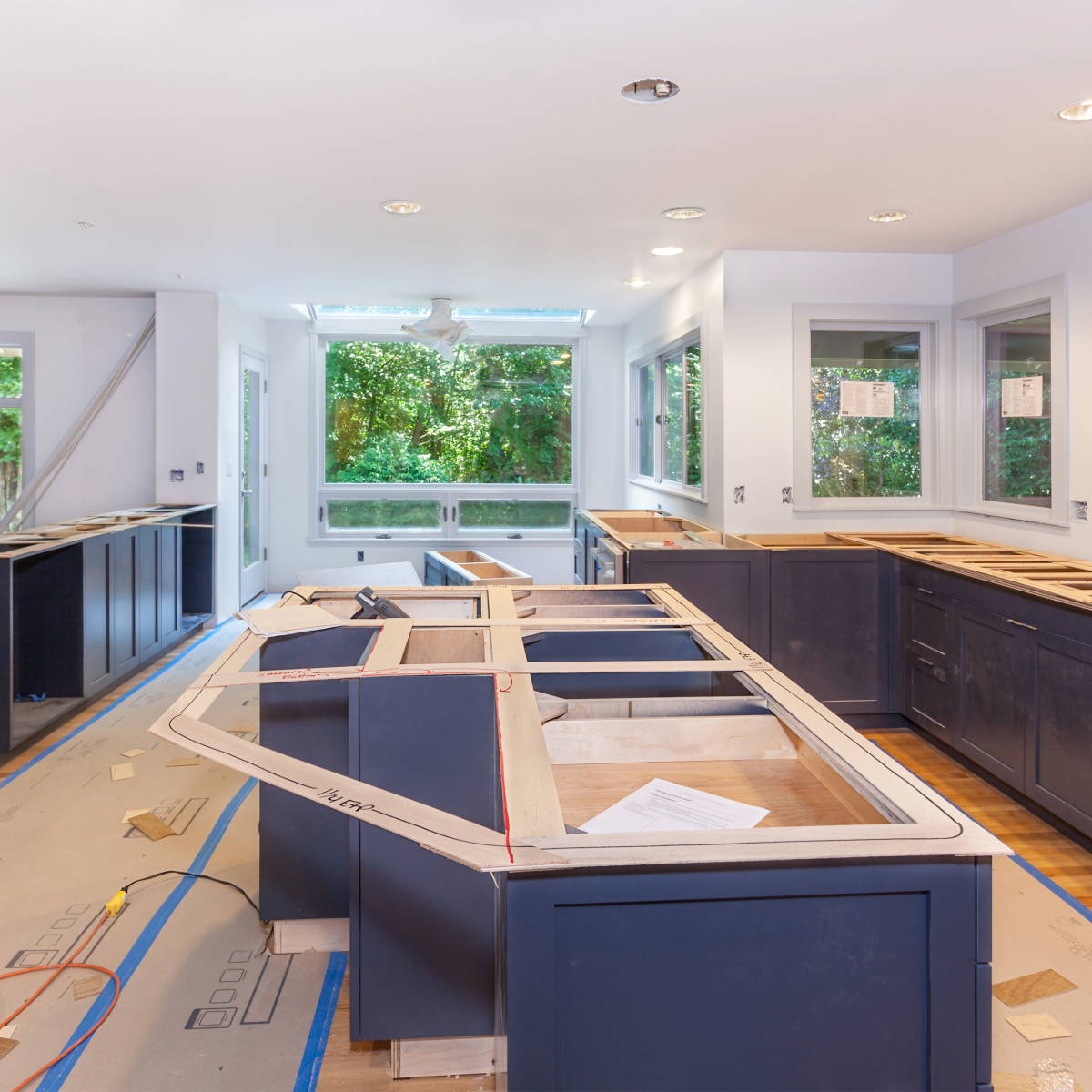 A spacious kitchen during renovation with dark blue cabinetry, a large central island, and windows overlooking greenery.