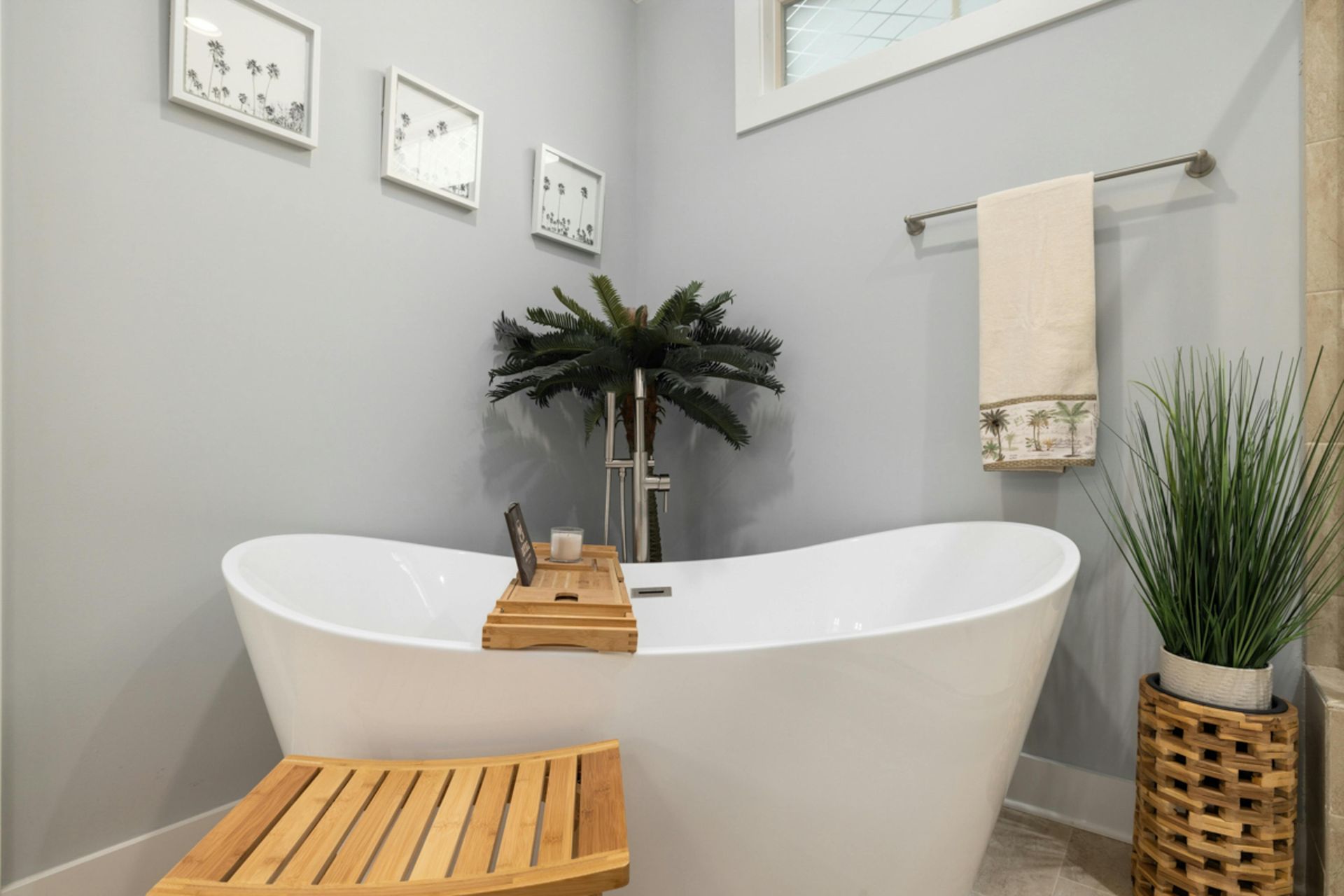 A freestanding white bathtub sits in a corner with gray walls, a wooden bath tray, a towel, and indoor plants.